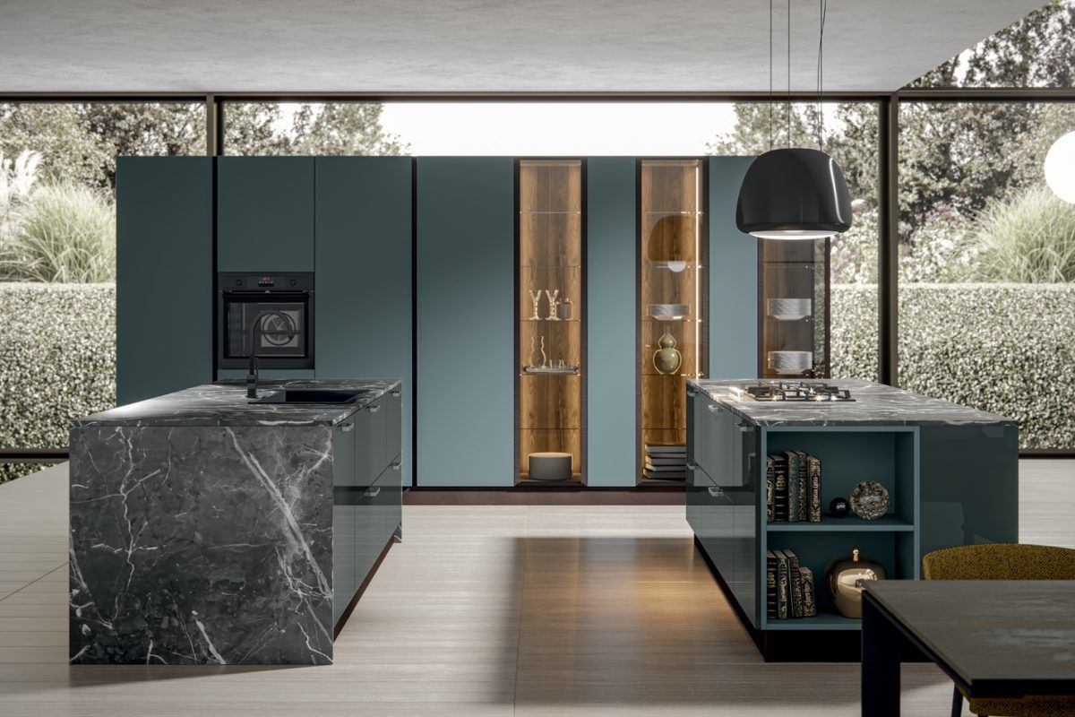 Cucina moderna con isola in marmo scuro, mobili color verde acqua e vani contenitori con ante in vetro in una stanza luminosa.