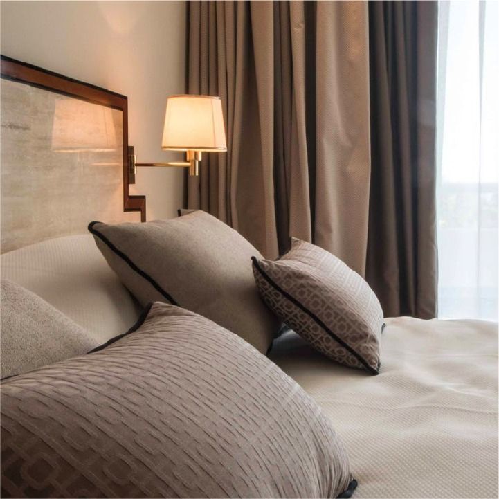Accogliente camera da letto con cuscini grigi, lenzuola beige, una lampada accesa e tende accanto a una finestra luminosa.