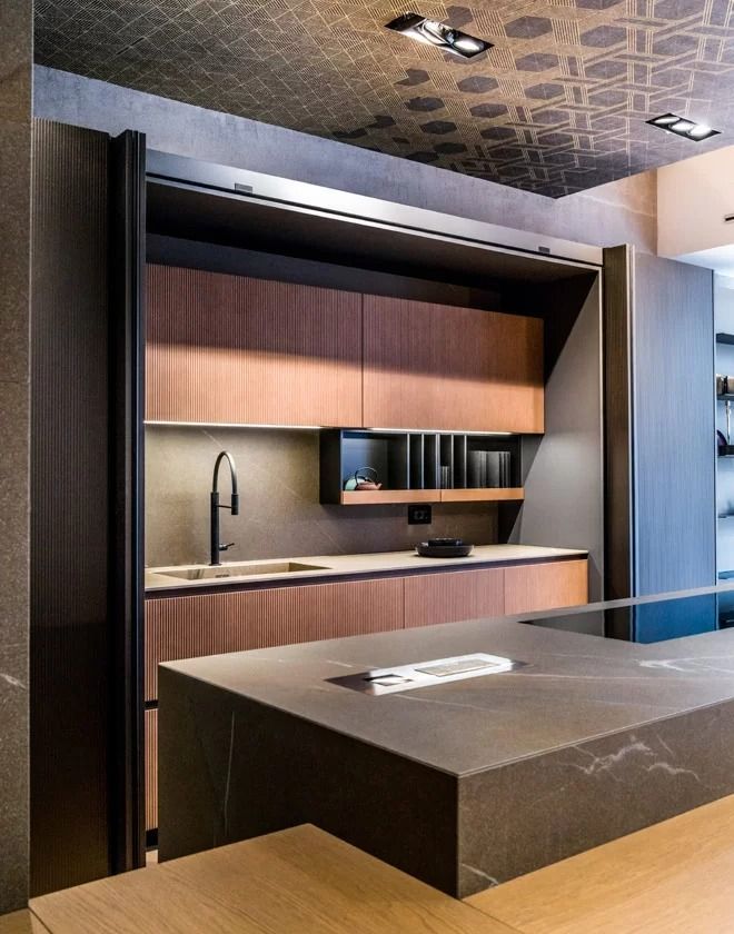 Cucina moderna con mobili in legno, piano di lavoro nero e isola centrale illuminata da faretti a soffitto con motivi decorativi.