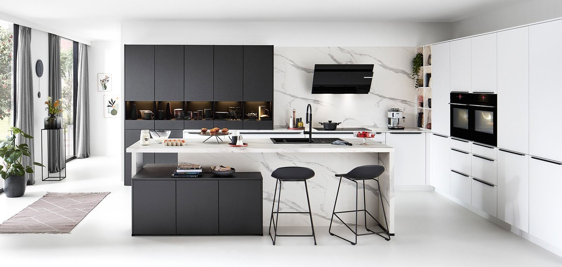 Cucina moderna bianca con dettagli neri, sgabelli sull'isola, forni a incasso e TV a parete.