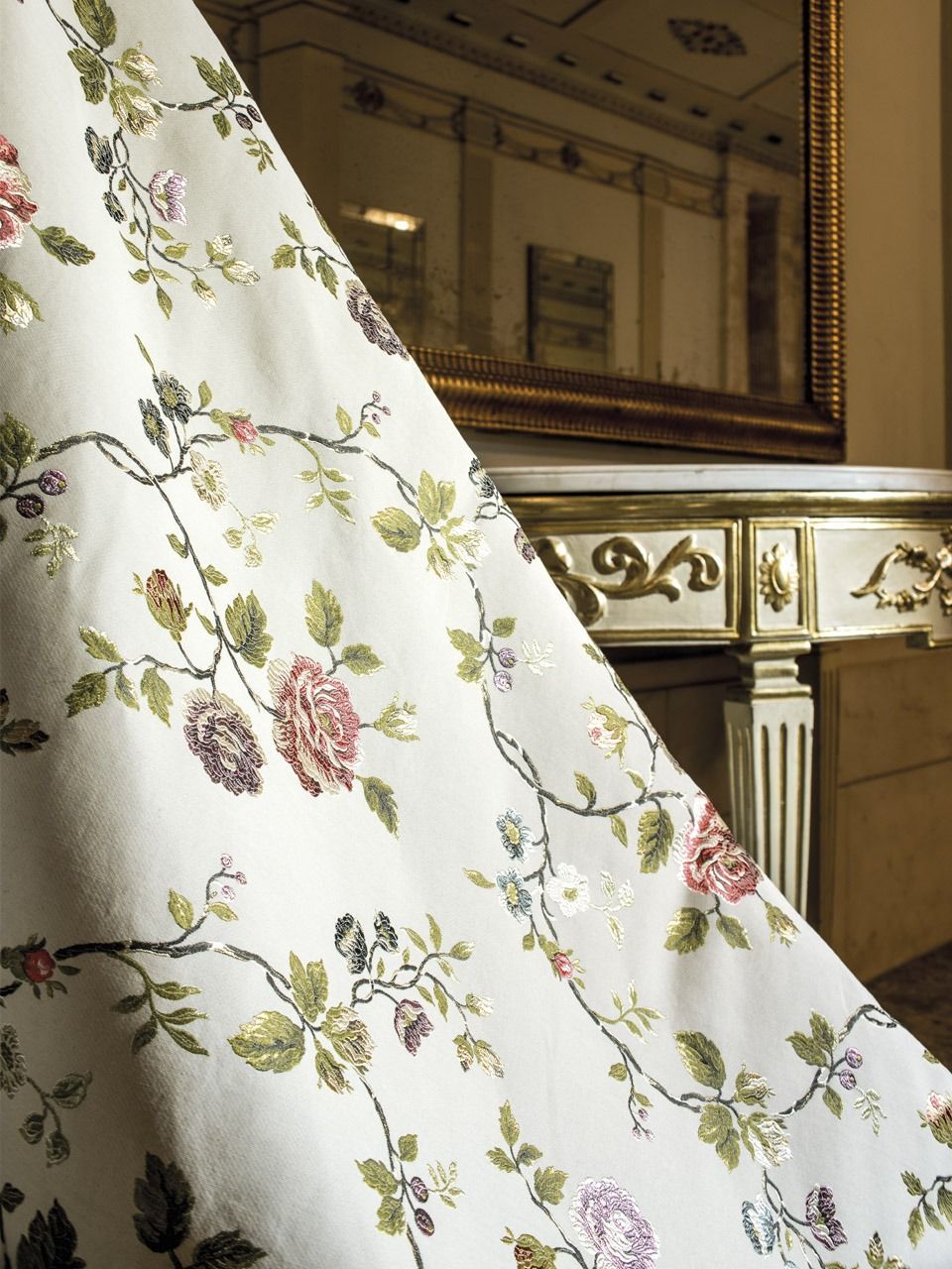 Tessuto bianco ricamato con motivi floreali drappeggiato davanti a un interno riccamente decorato con finiture dorate.