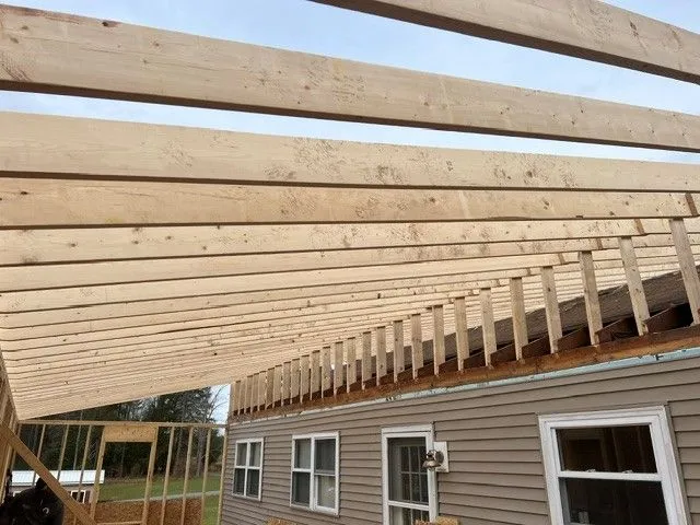Wooden roof rafters under construction beside a house, with exposed beams and wall framing.