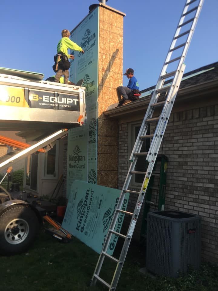 Two people installing green sheathing on a chimney exterior, one on a lift and the other on the roof.
