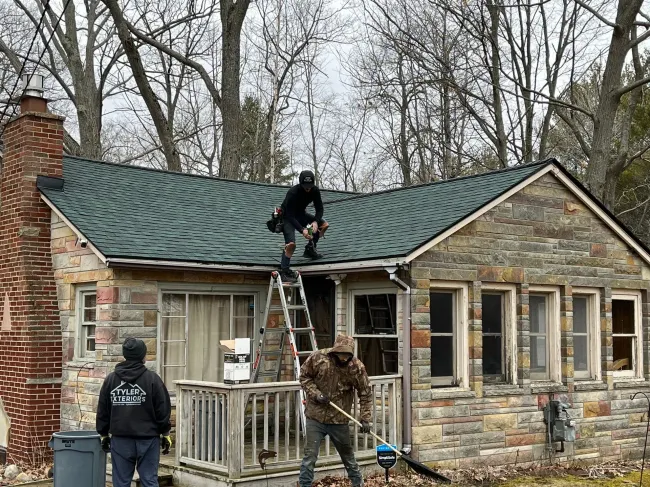 Workers on a roof of a stone house, one on a ladder. Overcast day.