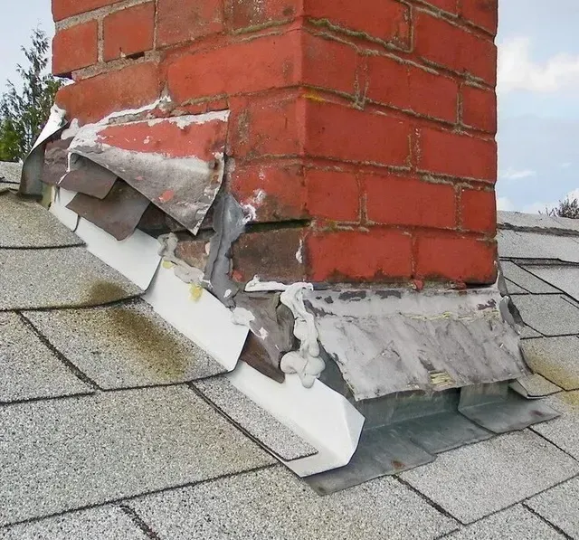 Brick chimney with damaged flashing on a gray shingled roof.
