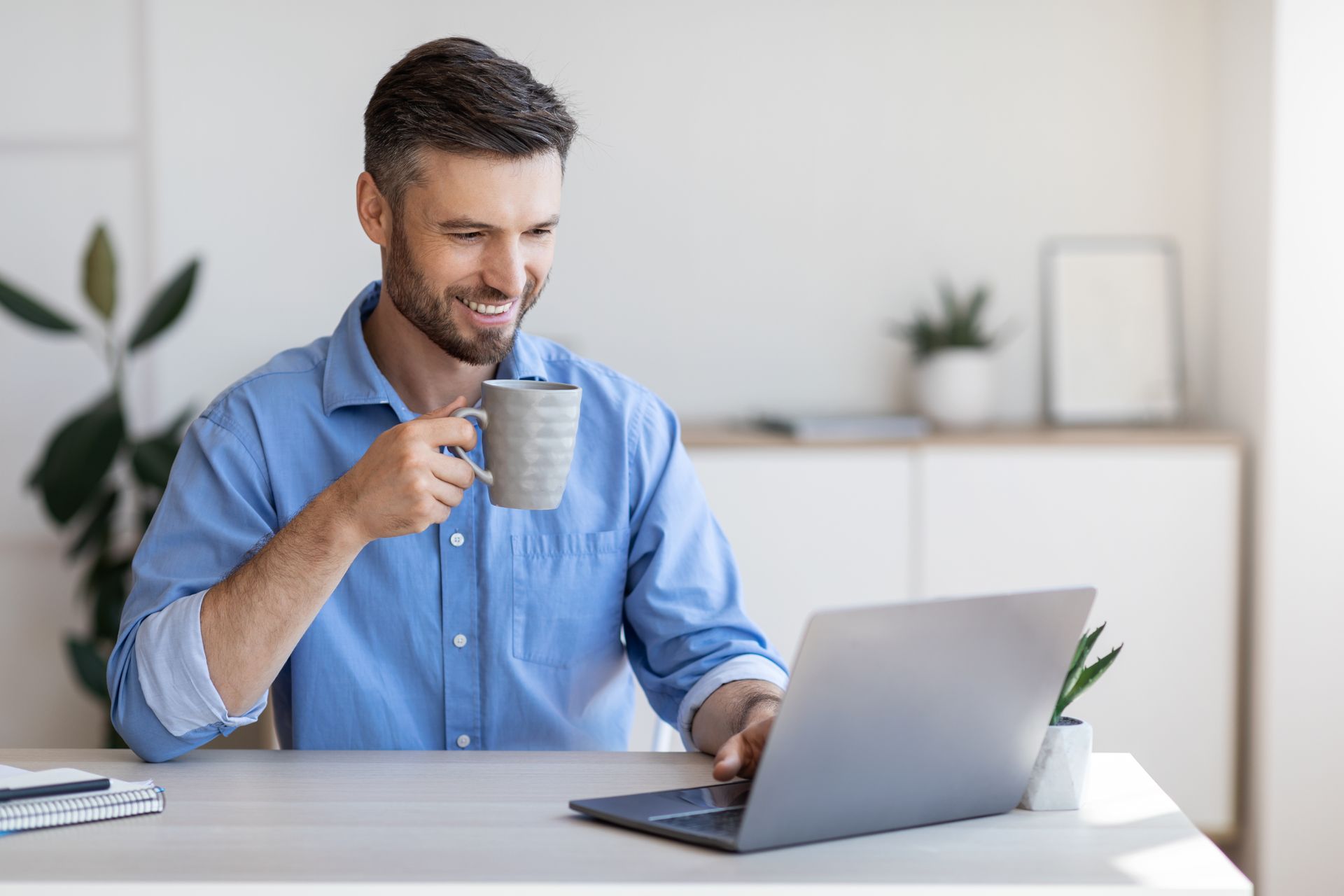 Man smiling, drinking coffee, working on laptop at desk in bright room.