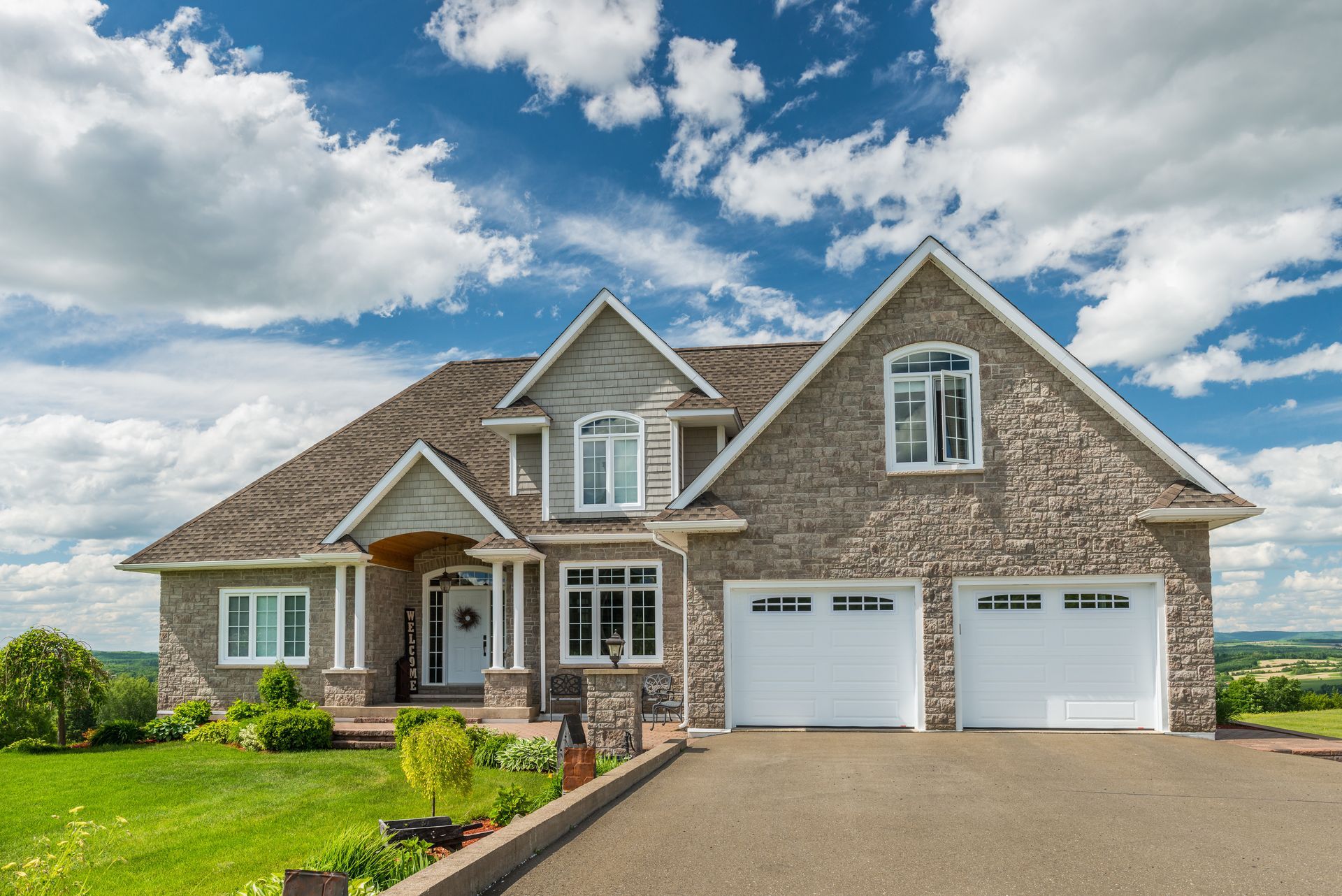 Stone-covered house with two-car garage, set on a green hill under a blue sky with fluffy clouds.