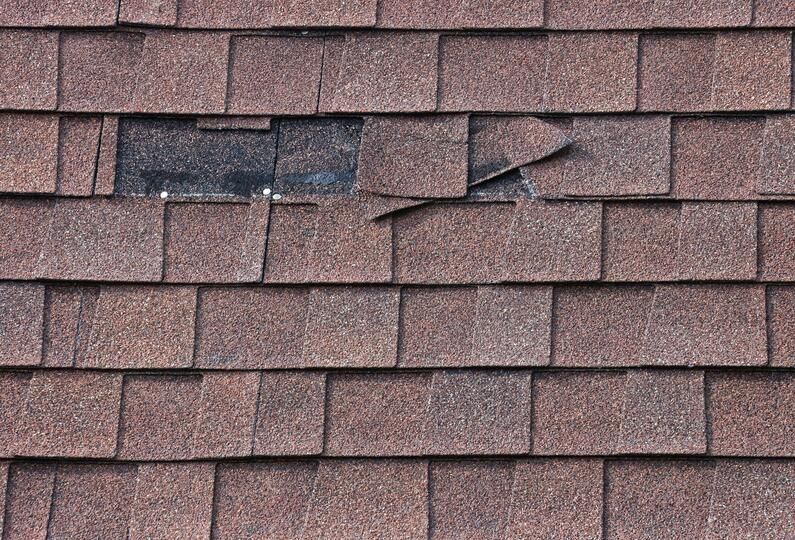 Close-up of a brown asphalt shingle roof with a missing section and damaged shingles.