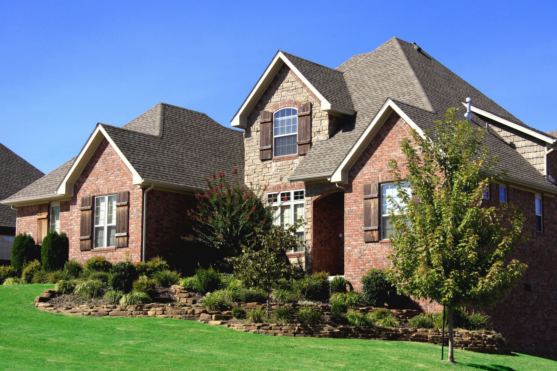 Brick house with a multi-level roof, stone accents, brown shutters, and manicured landscaping under a blue sky.