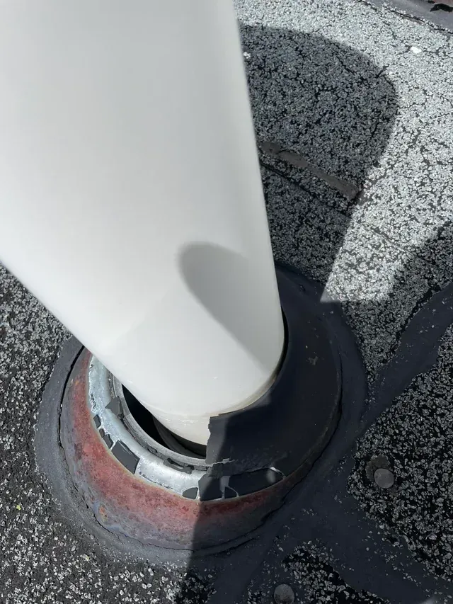 White pipe extending from a roof. The pipe is surrounded by a metal and rubber seal on a tar surface.