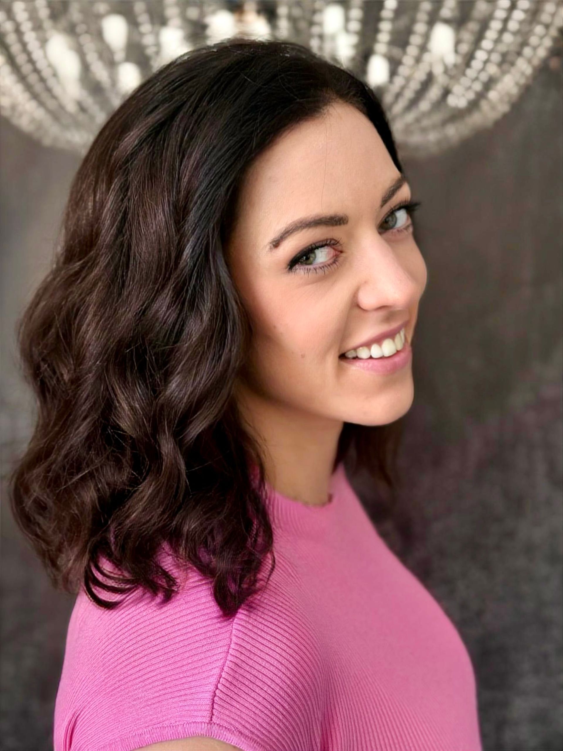 Eine Frau in einem rosa Pullover lächelt vor einem Kronleuchter.