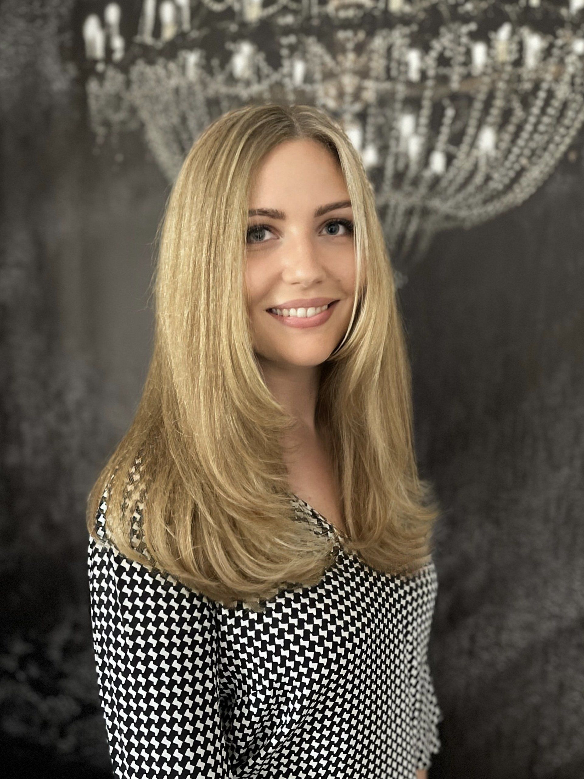 Eine Frau mit langen blonden Haaren lächelt vor einem Kronleuchter.