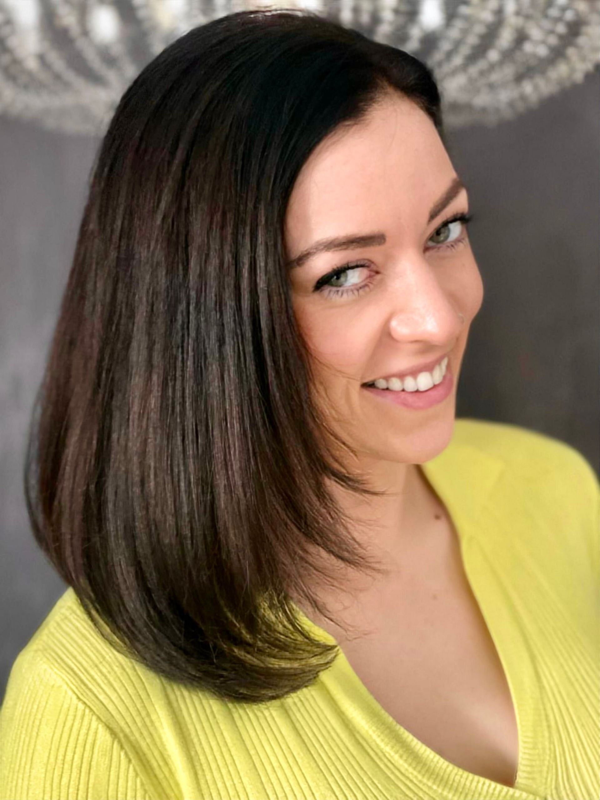 Eine Frau mit langen braunen Haaren trägt einen gelben Pullover und lächelt.