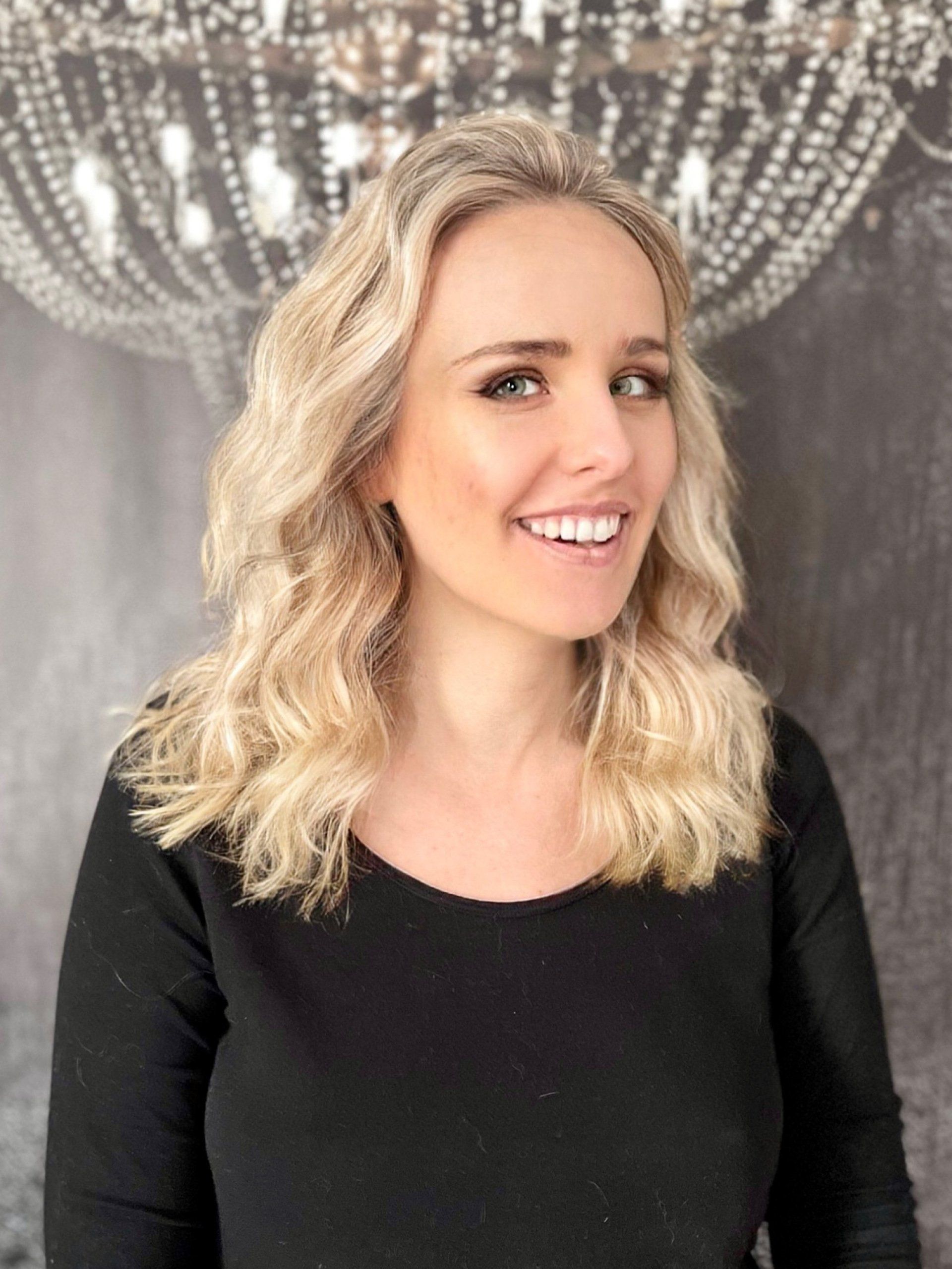 Eine Frau mit blondem Haar trägt ein schwarzes Hemd und lächelt vor einem Kronleuchter.