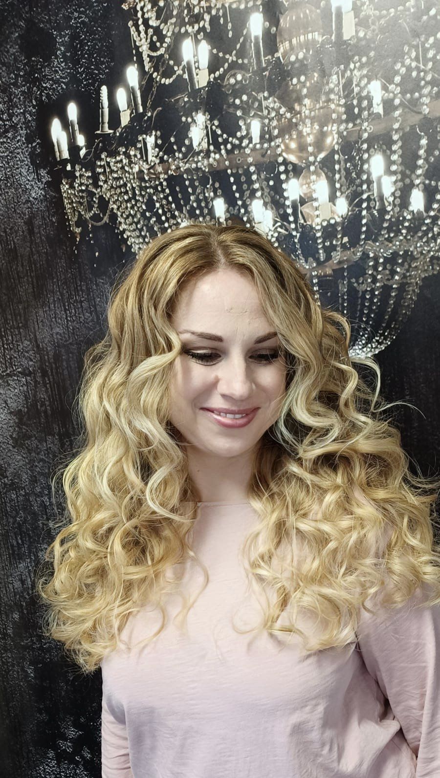 Eine Frau mit langen blonden Locken lächelt vor einem Kronleuchter.
