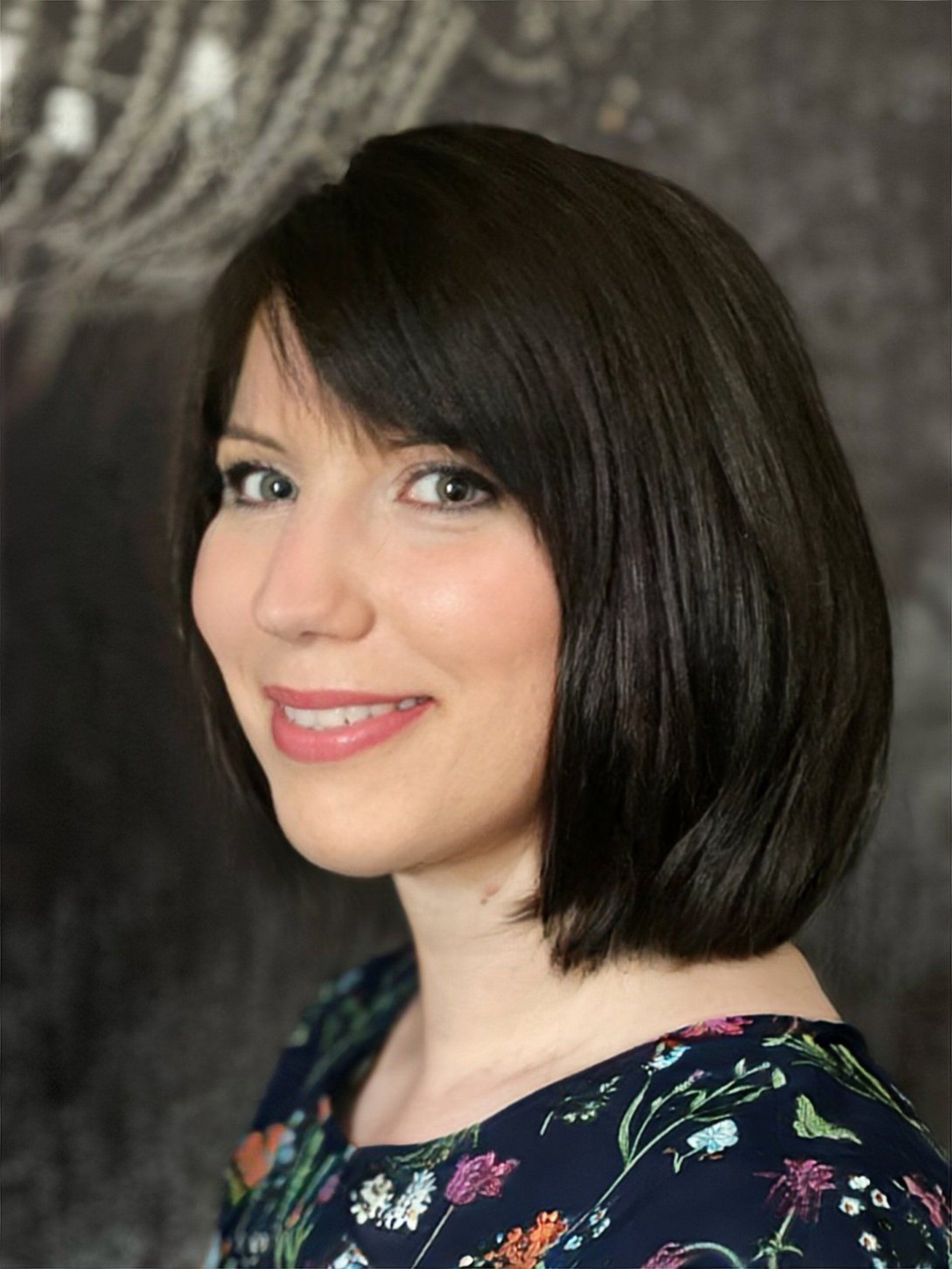 Eine Frau mit kurzen dunklen Haaren lächelt und trägt eine geblümte Bluse.