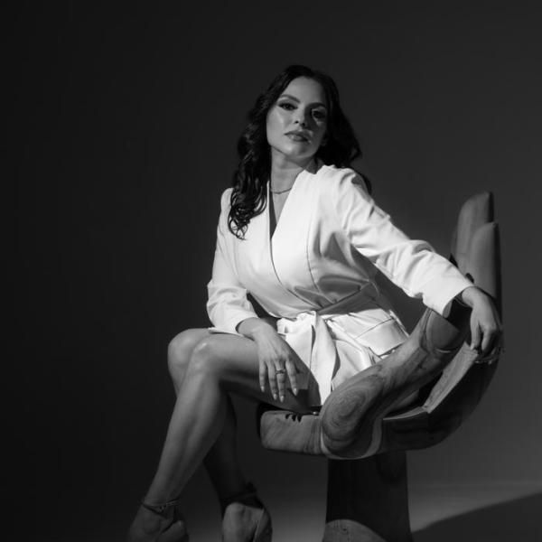 Dr Kaly Castellanos sits on a sculptural wooden chair against a plain, dark background.