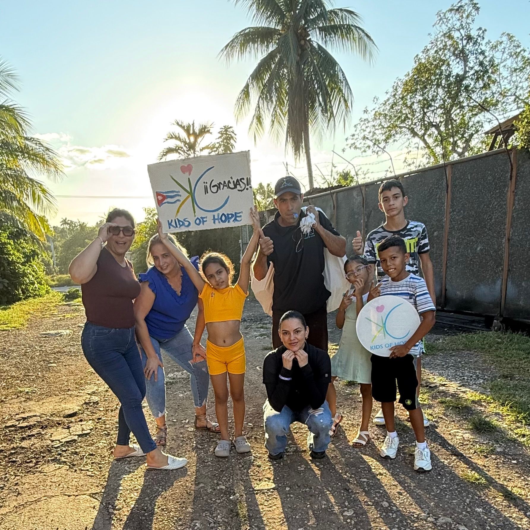 A group of people standing together outdoors on a dirt road, holding signs with text and graphics under a palm tree.