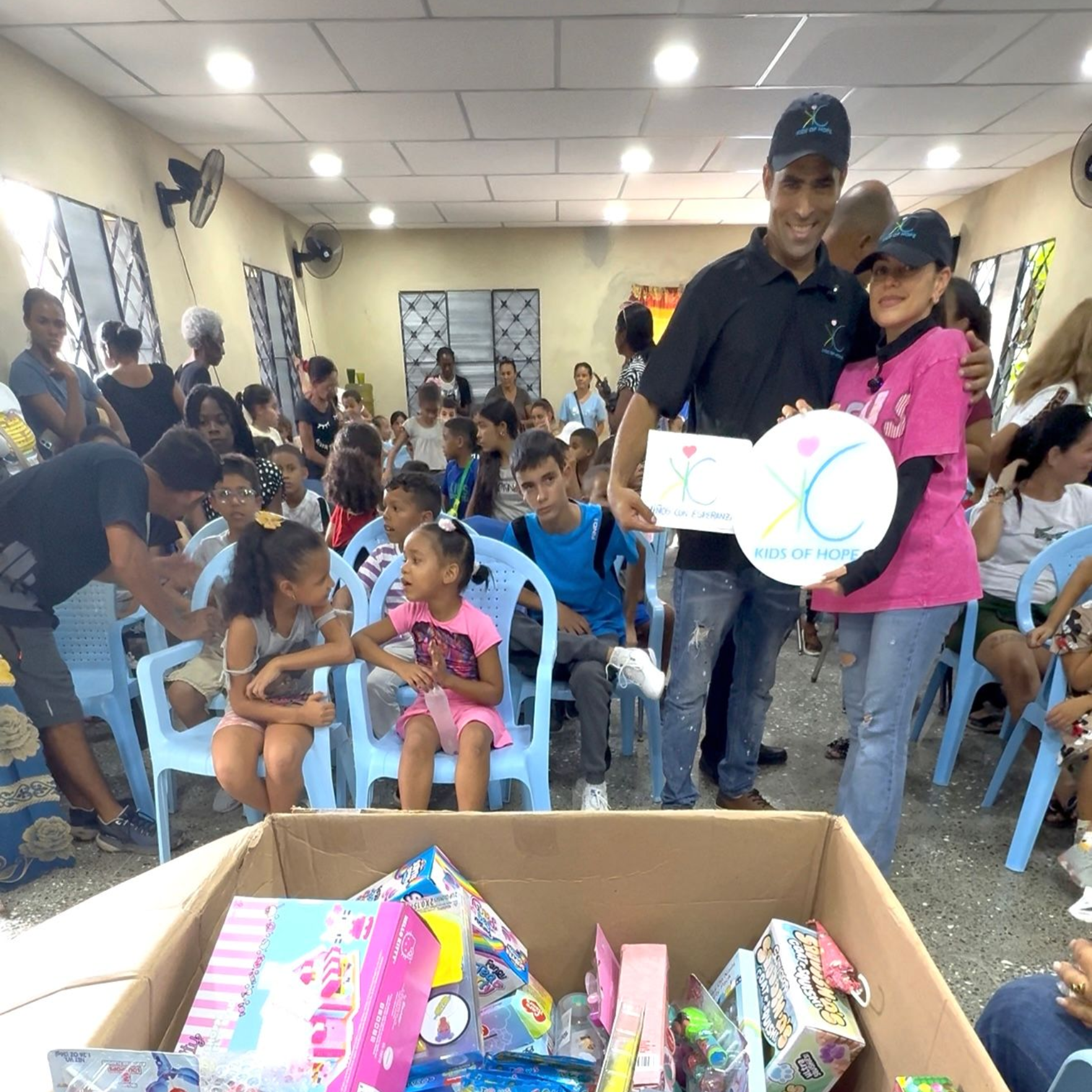 Two people hold a circular sign with a logo in a room filled with children; a cardboard box with gifts sits in front.
