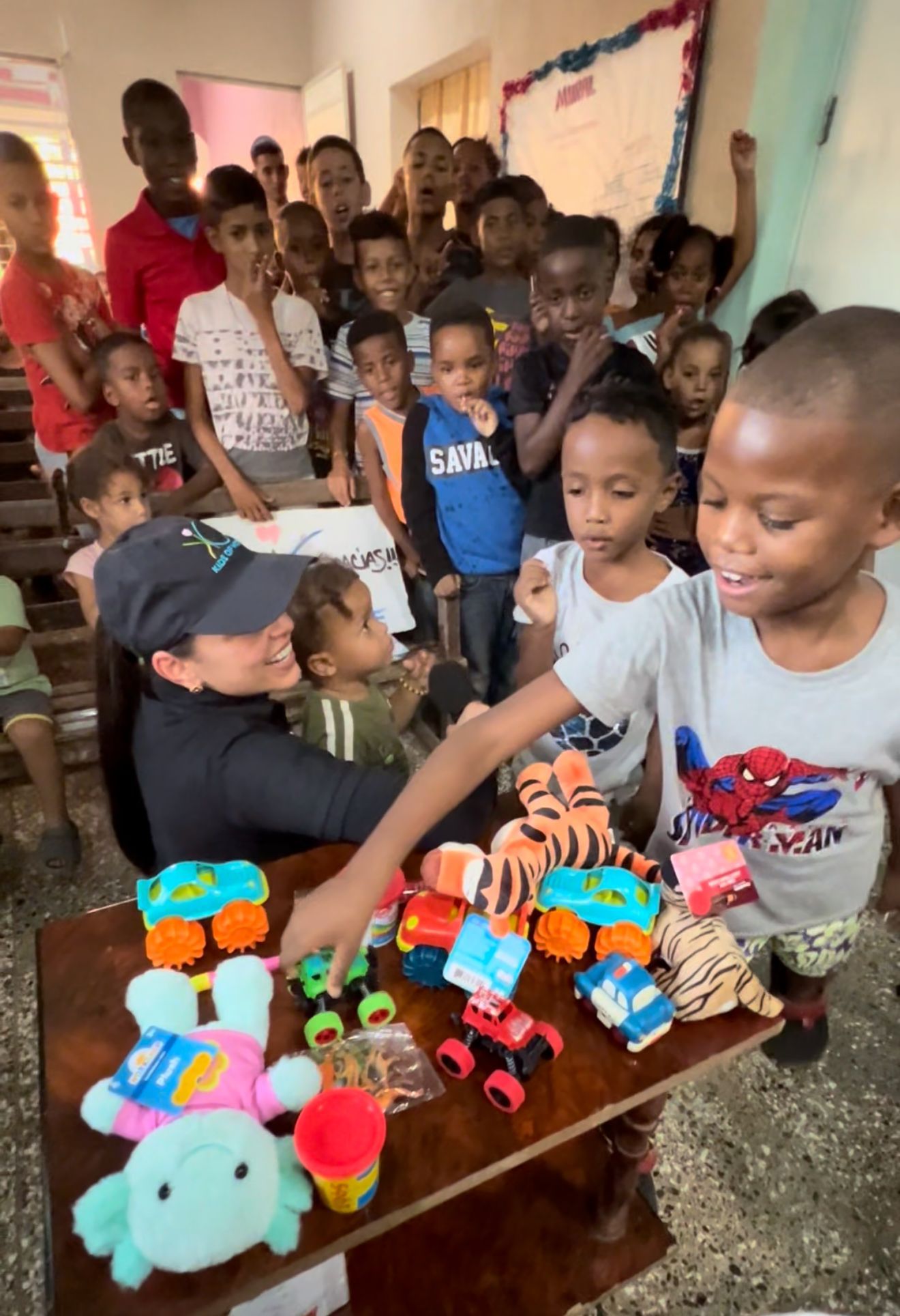 A person sitting at a table with toys, surrounded by a group of children selecting items in a classroom setting.