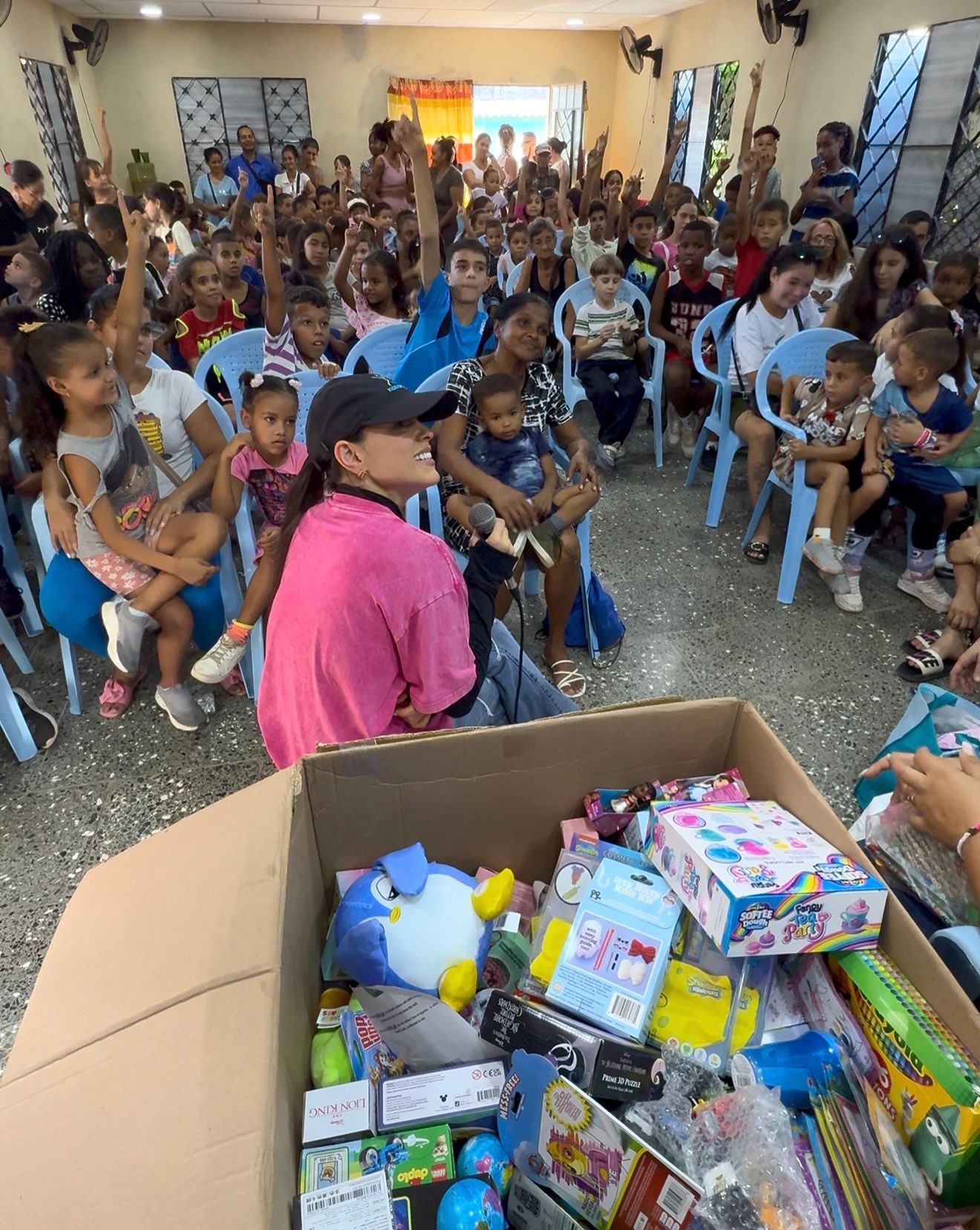 A person speaks to a crowded room of children, with a box of toys and supplies in the foreground.