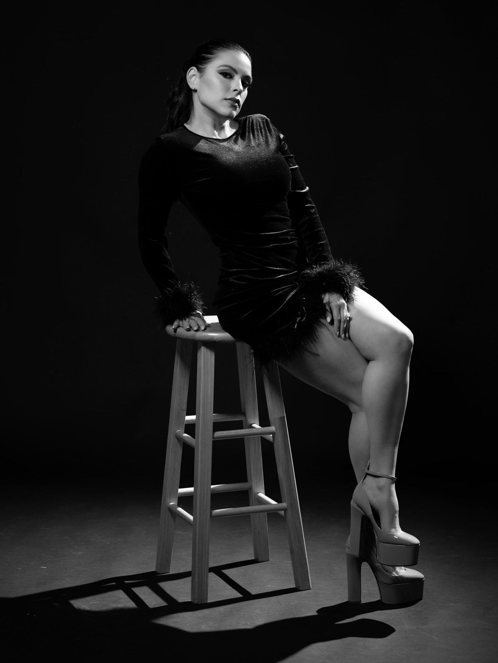 A person in a dark, feathered dress poses on a wooden stool against a black background, wearing high-platform heels.