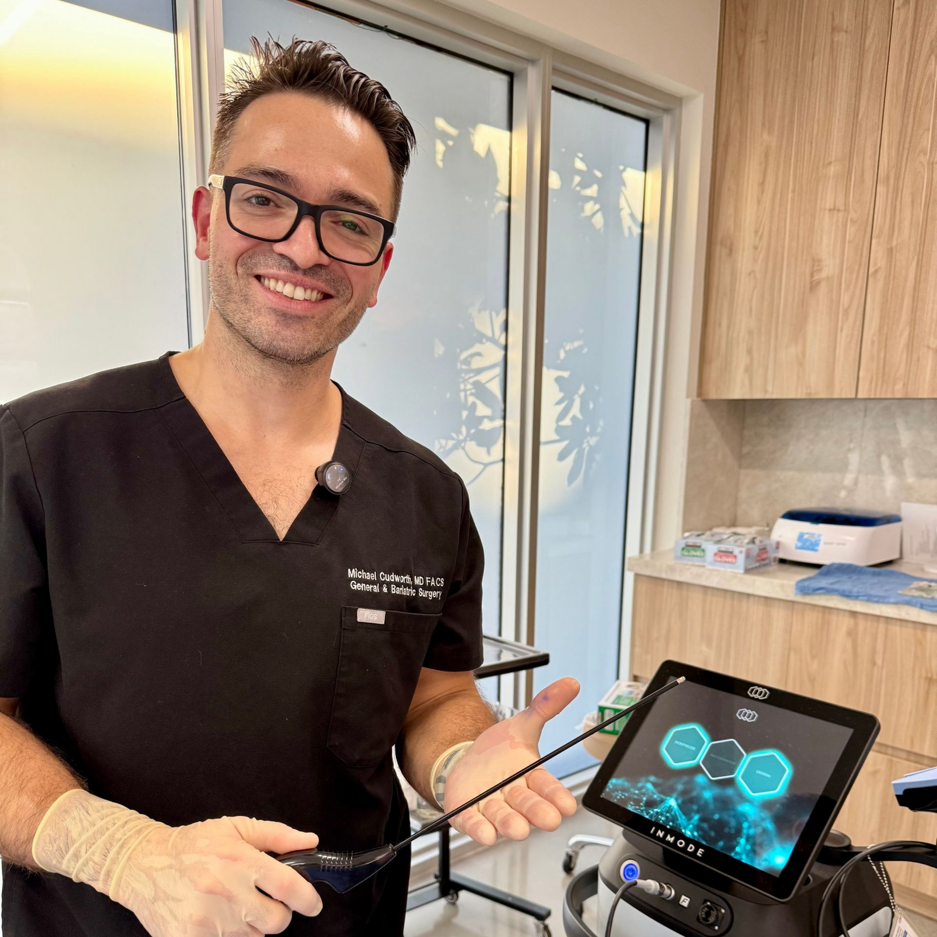 A smiling medical professional in black scrubs holds a handheld laser device next to a digital display screen.