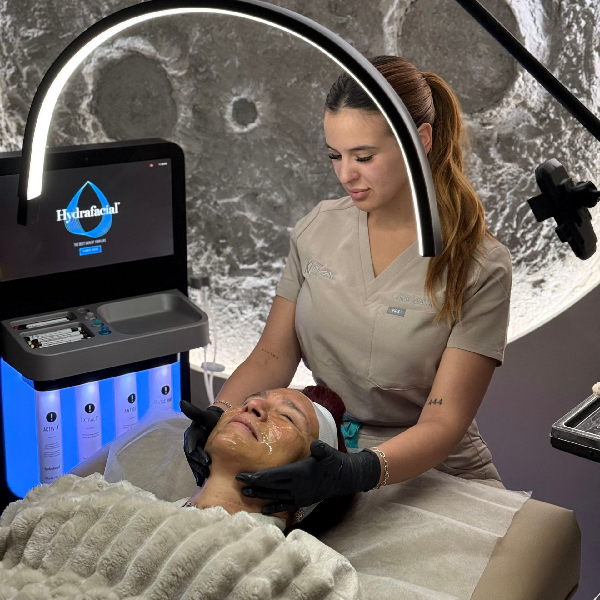 An aesthetician in scrubs and black gloves performs a facial treatment on a client using a HydraFacial machine.
