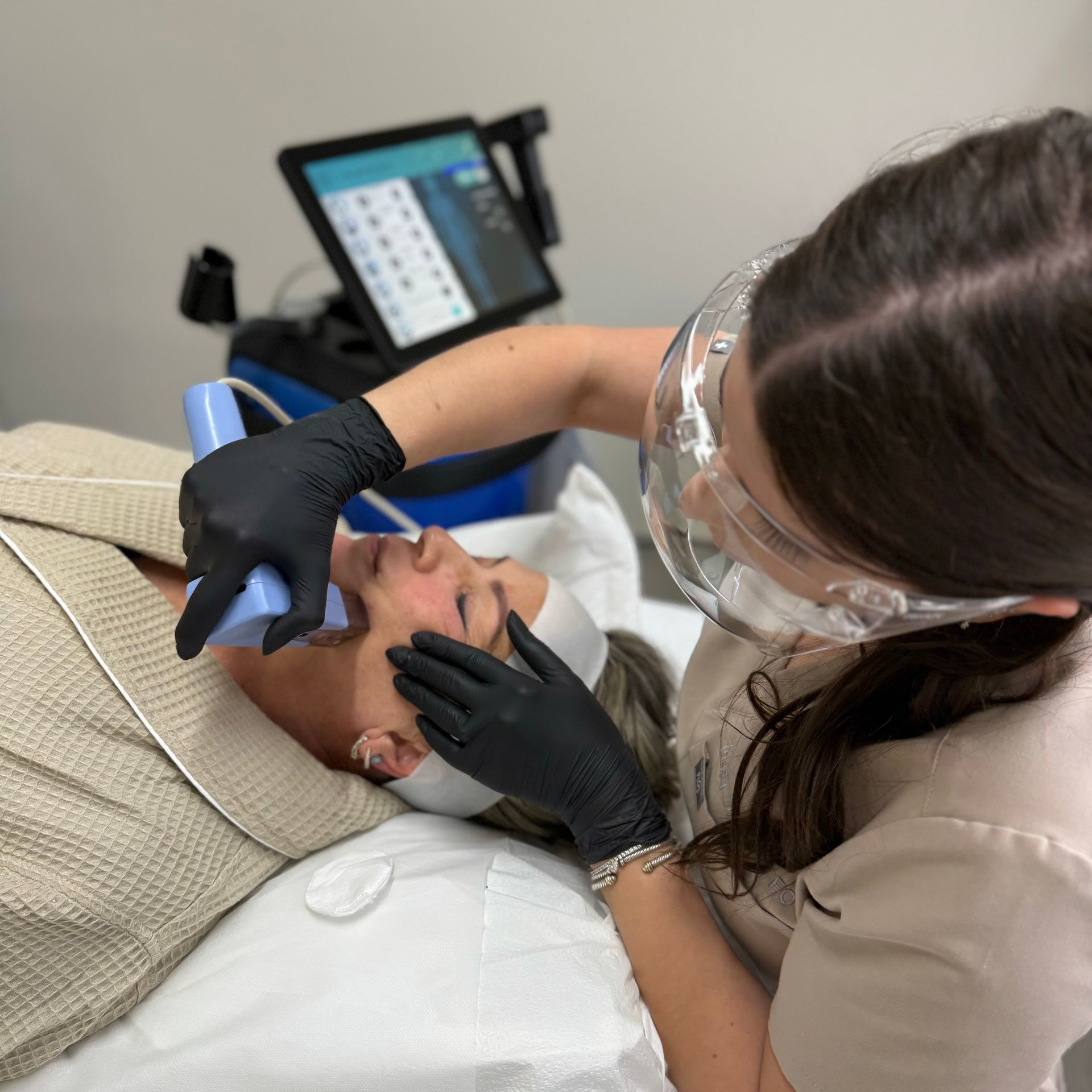 An aesthetician in protective eyewear and black gloves uses a handheld device on a client’s face during a skin treatment.