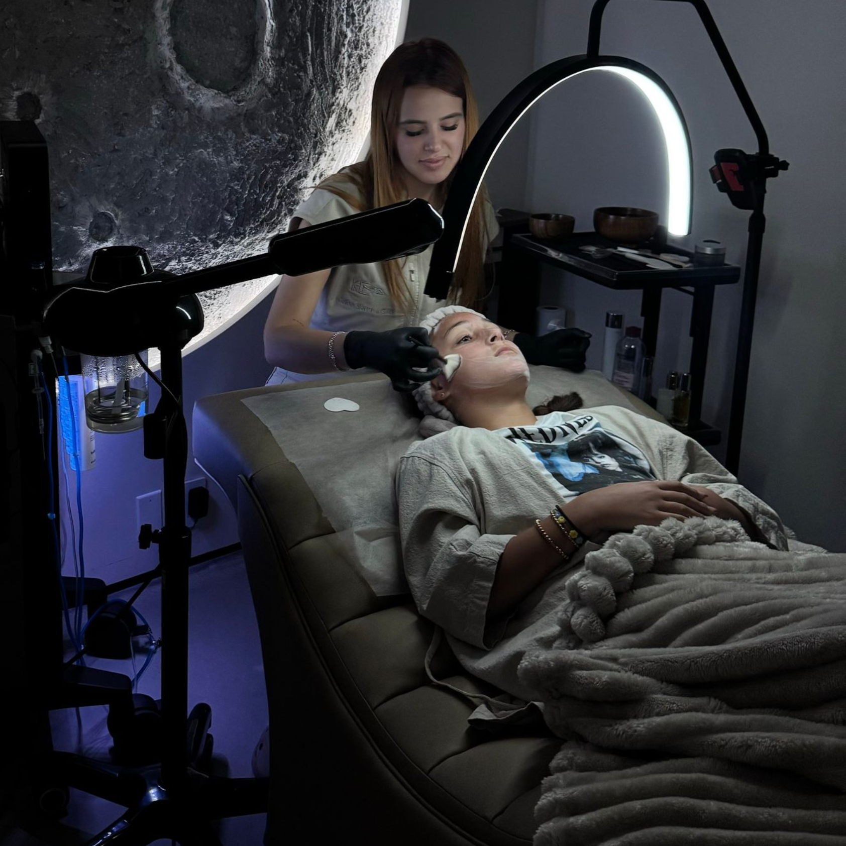 An esthetician in black gloves performs a facial treatment on a person lying on a spa bed under a glowing ring light.