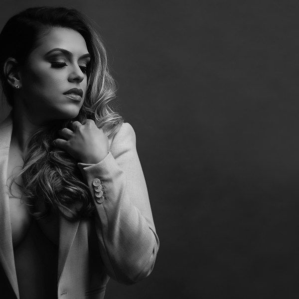 A black-and-white portrait of a person wearing a light blazer, looking down with a hand raised toward their hair.