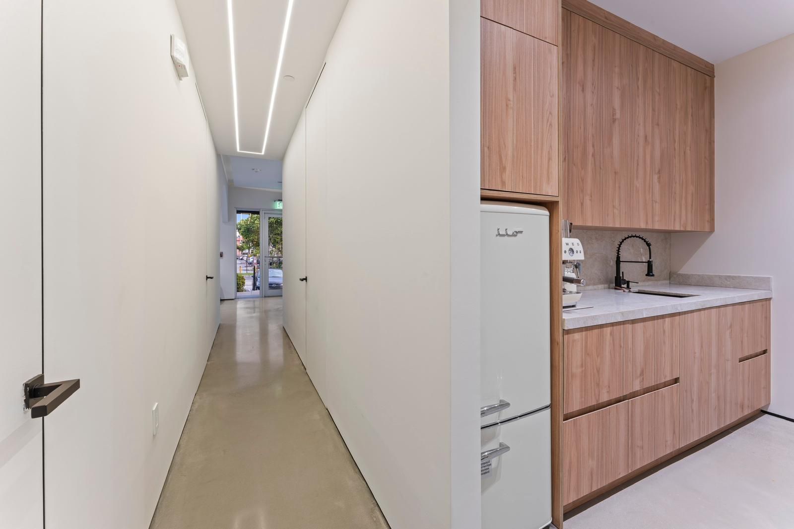 A hallway with linear ceiling lighting leads to an exterior door, next to a modern kitchen with light wood cabinetry.