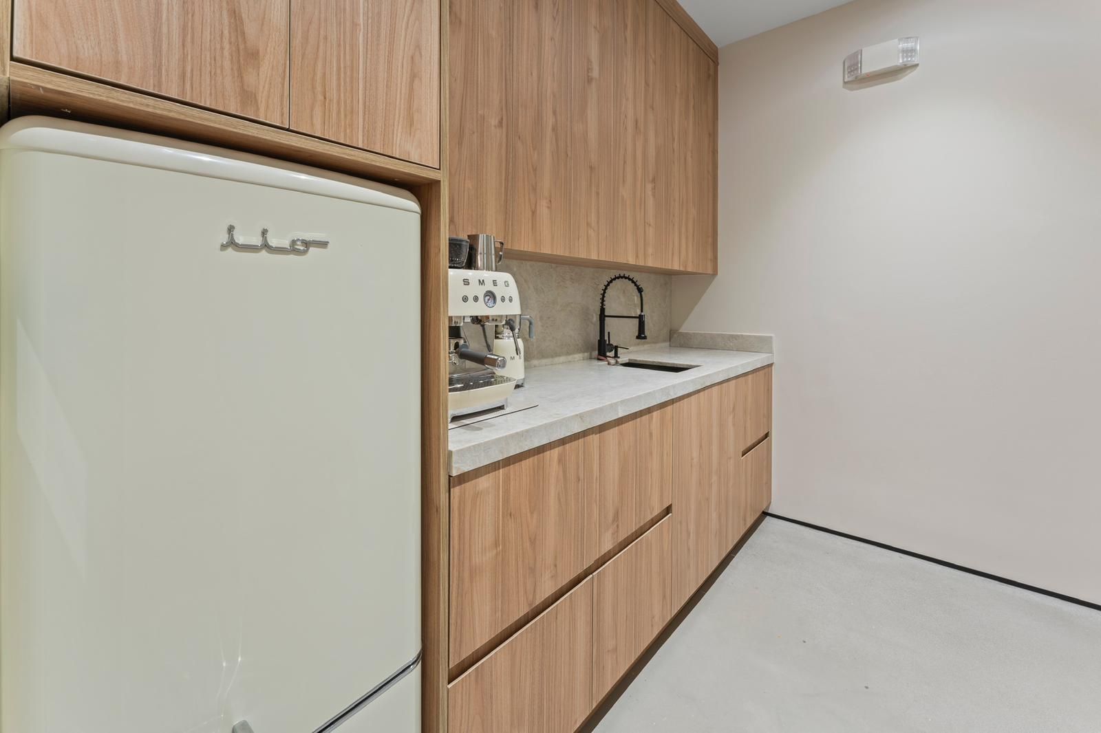 A modern kitchen features a cream-colored refrigerator, light wood cabinetry, a white countertop, and a coffee machine.