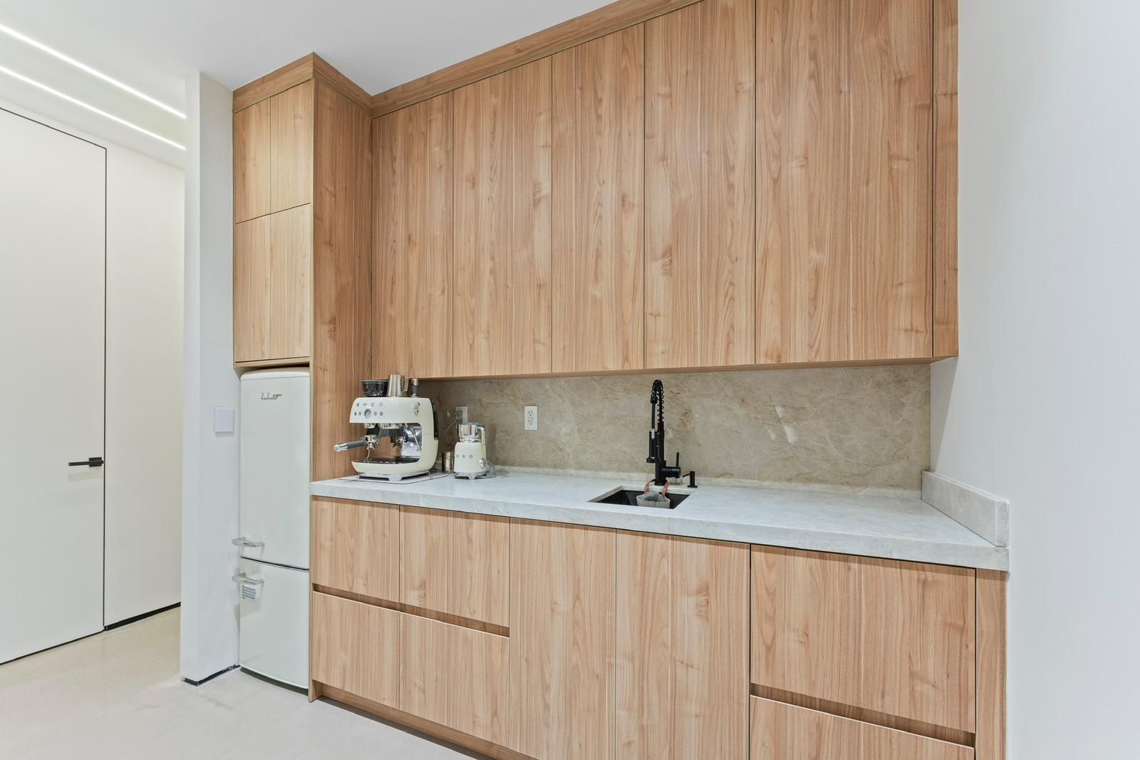 A modern kitchen features light wood cabinets, white countertops, a small sink, and an espresso machine on the counter.