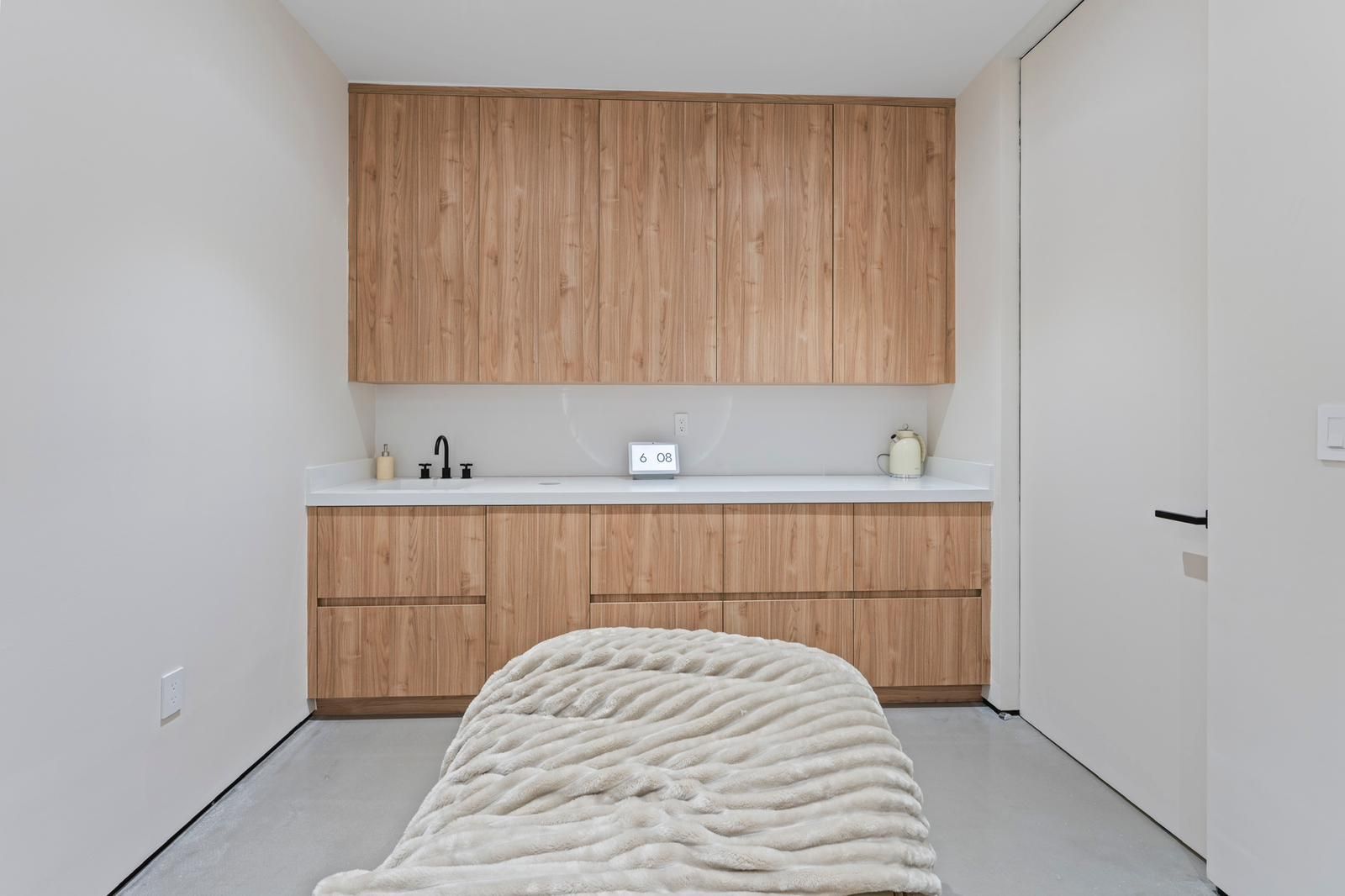 A treatment room with light wood cabinets, a white countertop with a sink, and a textured cream massage table.