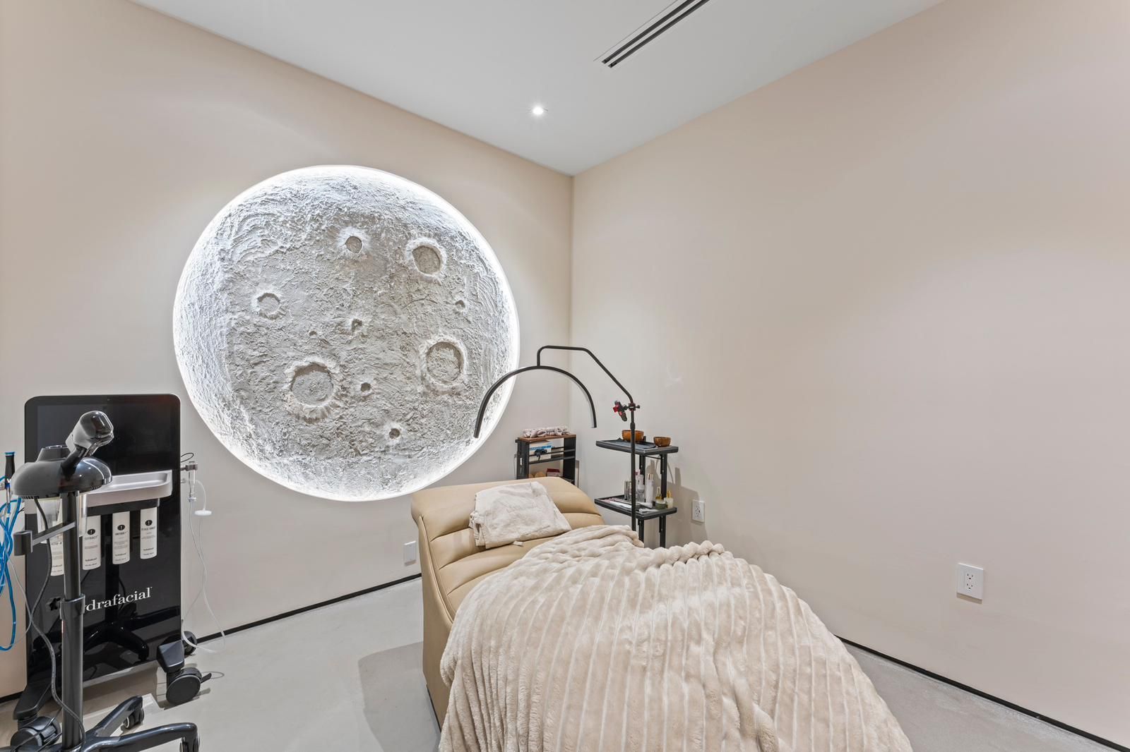 A spa treatment room featuring a round, glowing moon wall art above a beige massage table with a white textured throw.