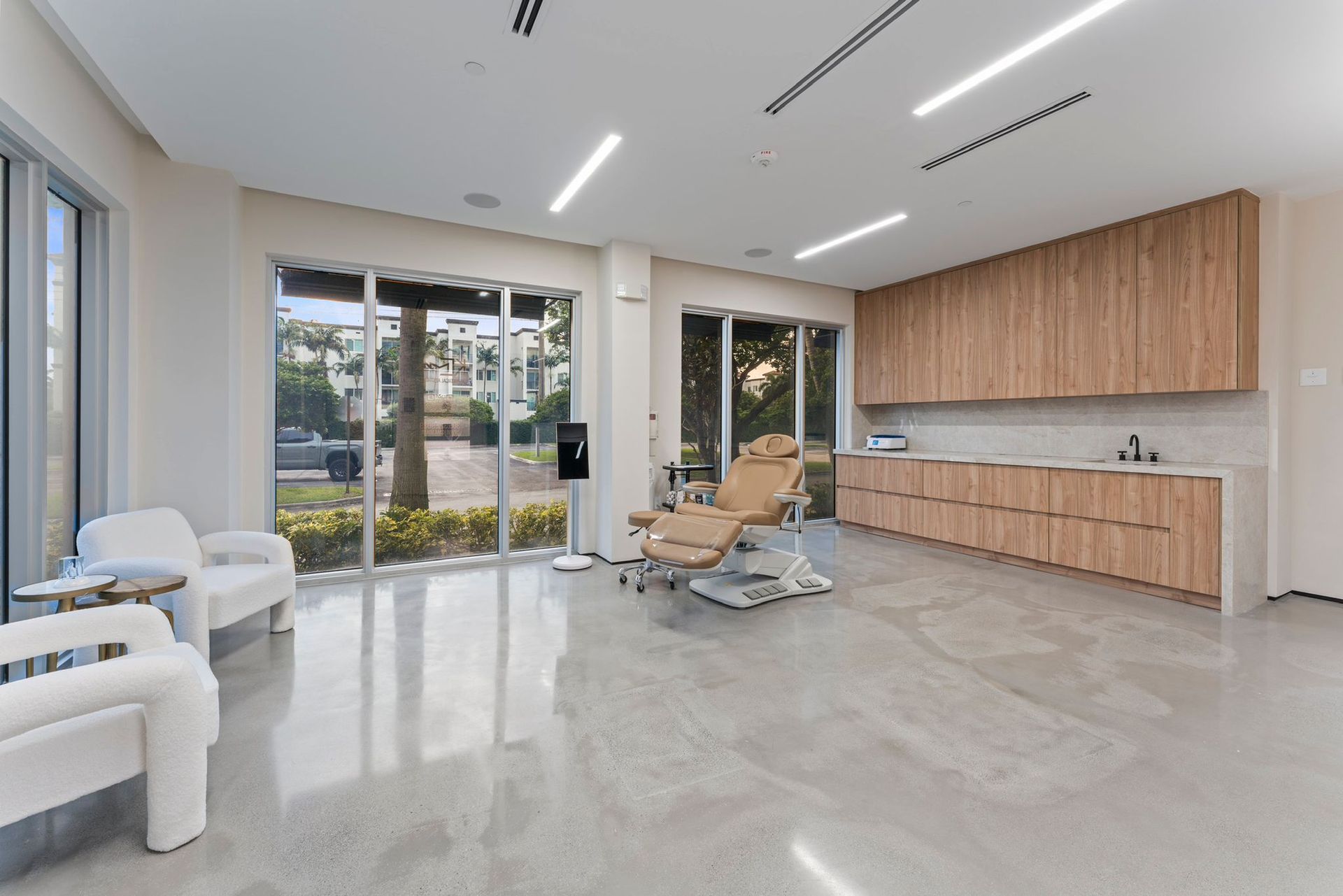 A bright, modern medical spa room with white chairs, a treatment bed, and light wood cabinetry against a beige wall.