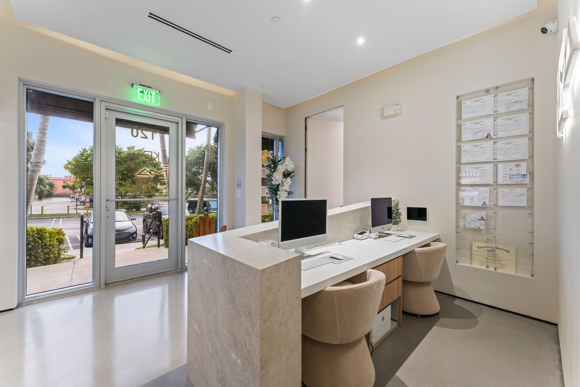 A modern reception area with a marble desk, two computers, and chairs, located near a glass entrance door.