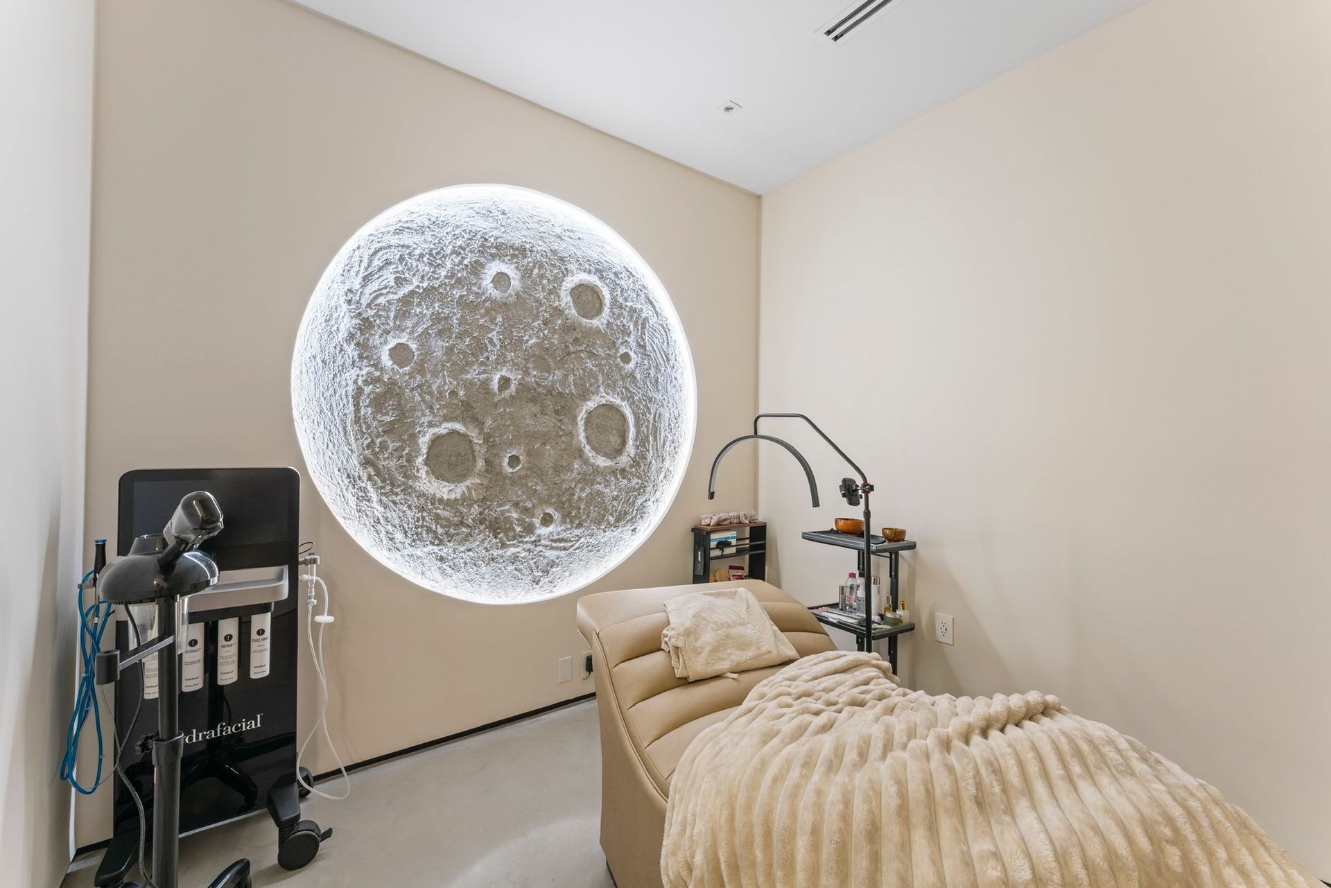 A spa treatment room with a beige massage bed, a moon-themed wall light, and professional skincare equipment.