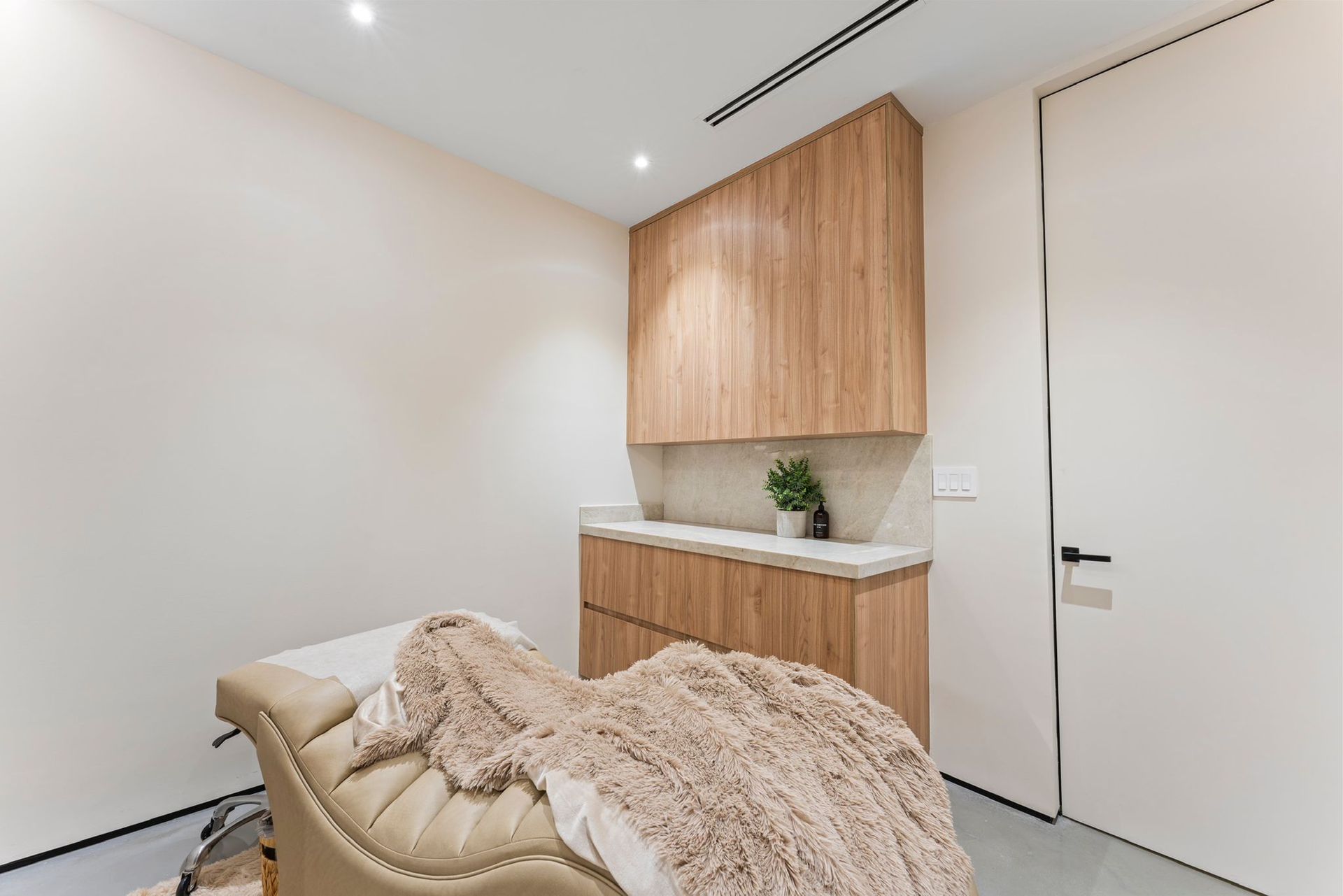 A treatment room with a beige massage bed, a textured blanket, and light wood cabinets against a white wall.