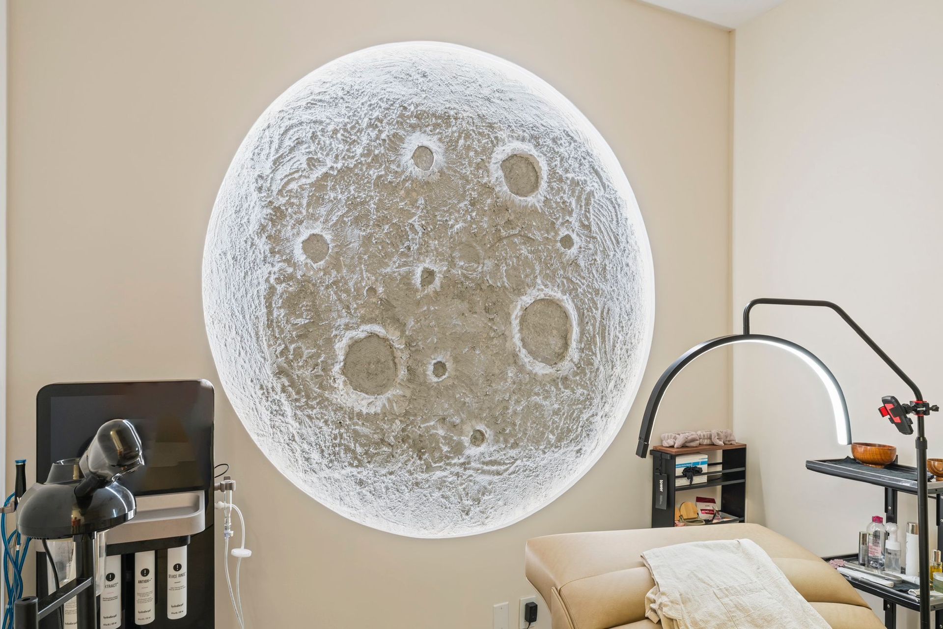 A large, round, textured moon mural glows on a tan wall in an esthetician's treatment room with medical equipment nearby.