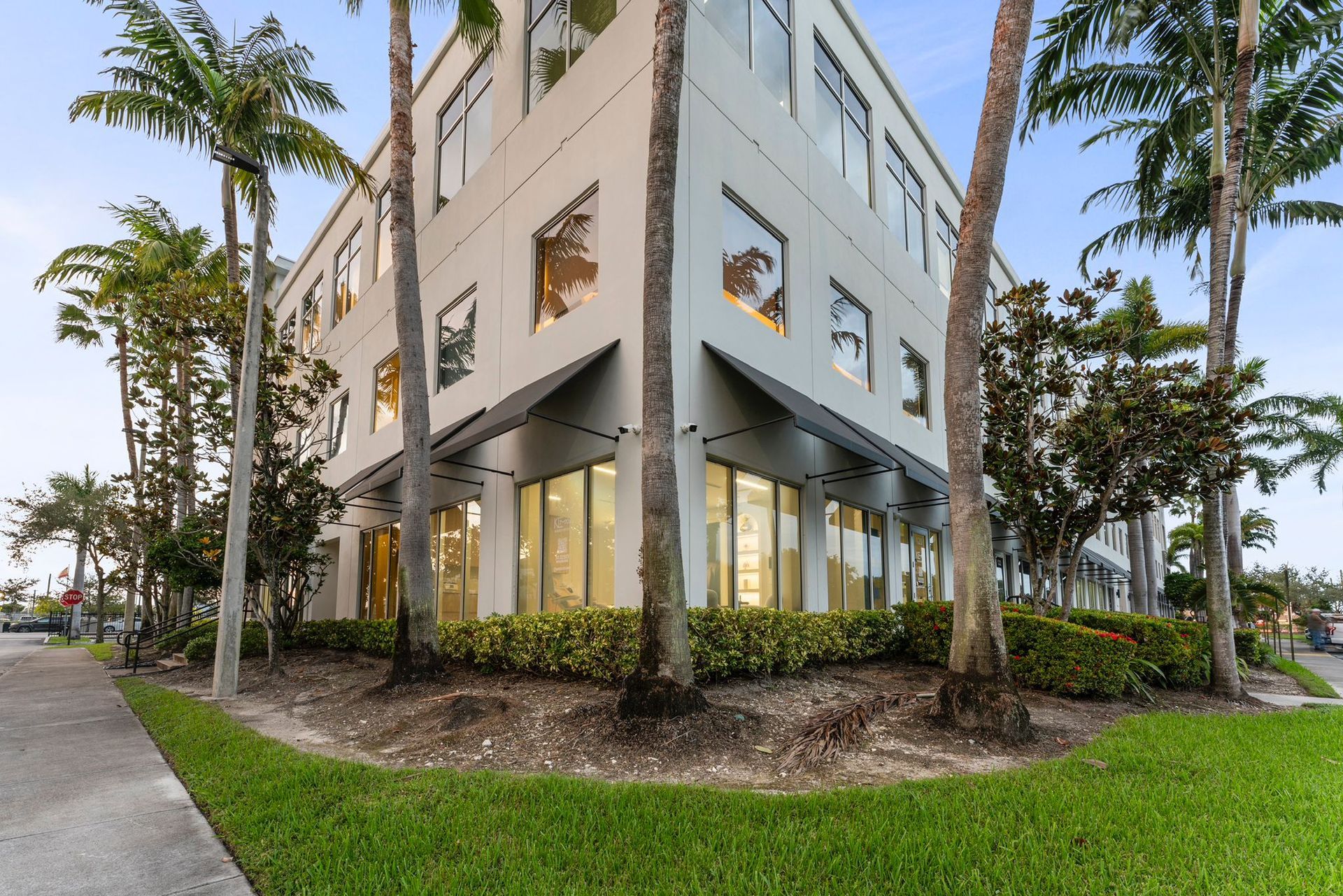 A multi-story beige office building with dark window awnings, surrounded by palm trees and landscaped hedges.