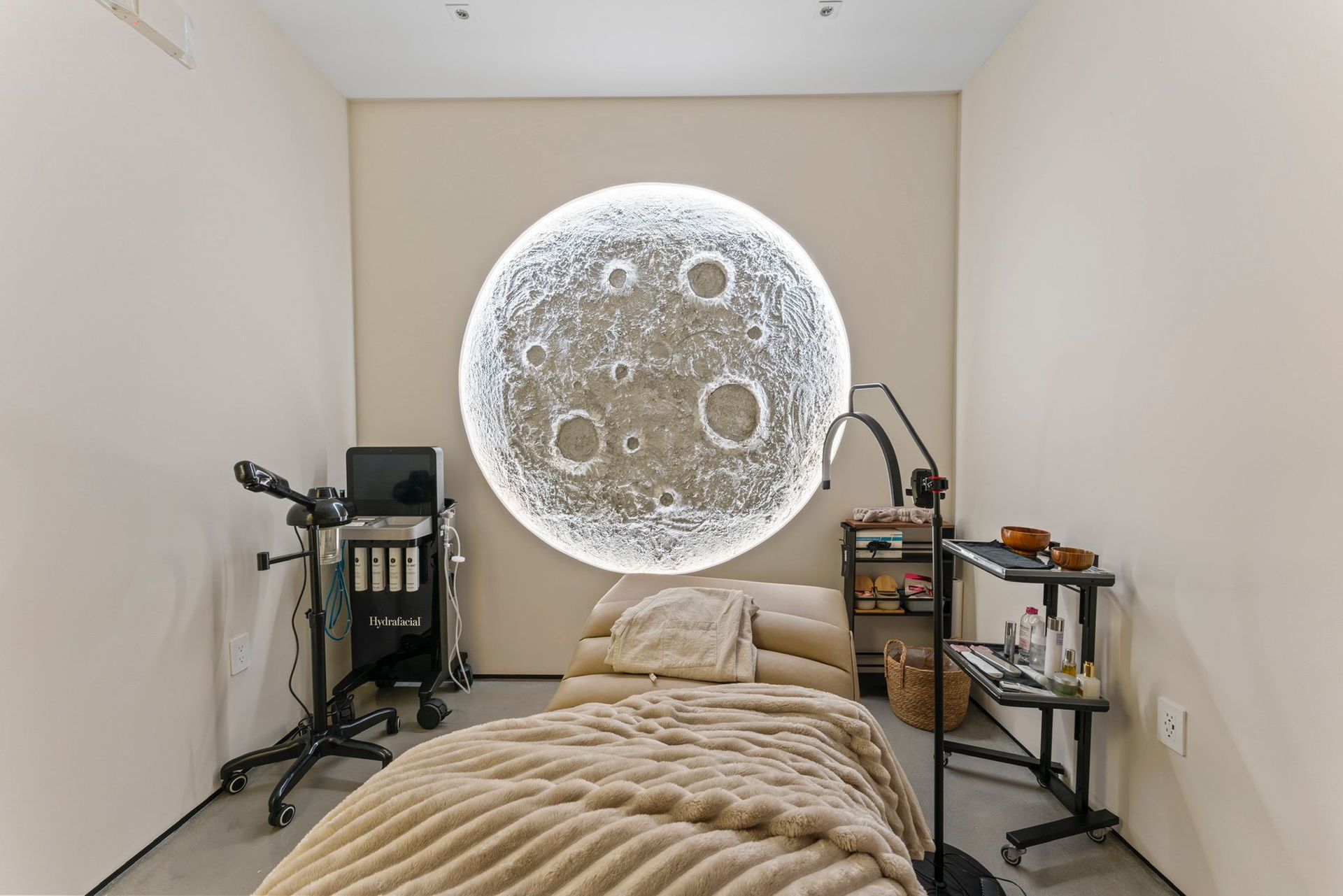 A neutral-toned treatment room featuring a round, illuminated moon wall art behind a beige massage table with equipment.