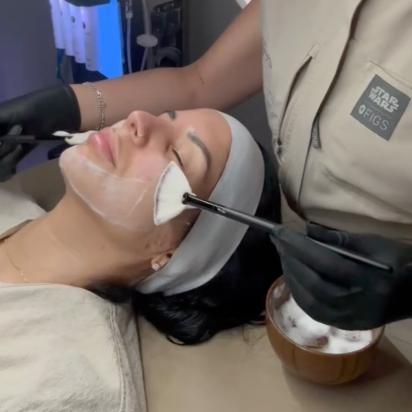 An aesthetician in a tan uniform applies a white facial mask to a client's cheek using a fan brush.