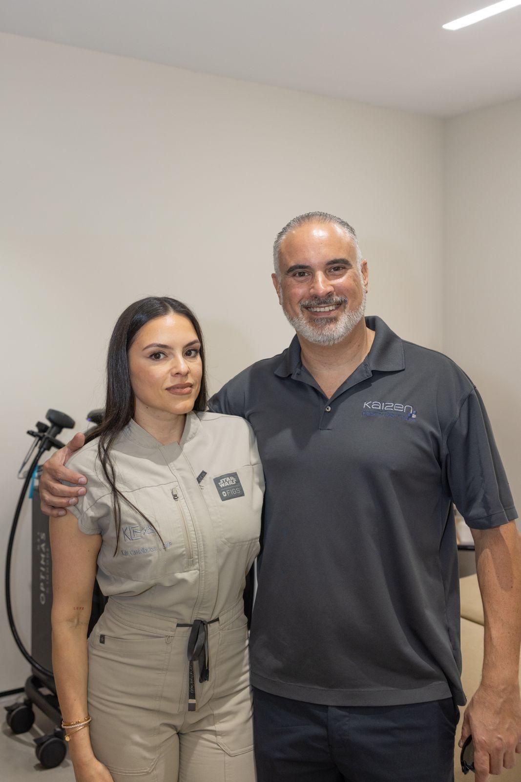 A man and a woman in uniforms stand side-by-side in a medical office, smiling for a photo with the man's arm around her.