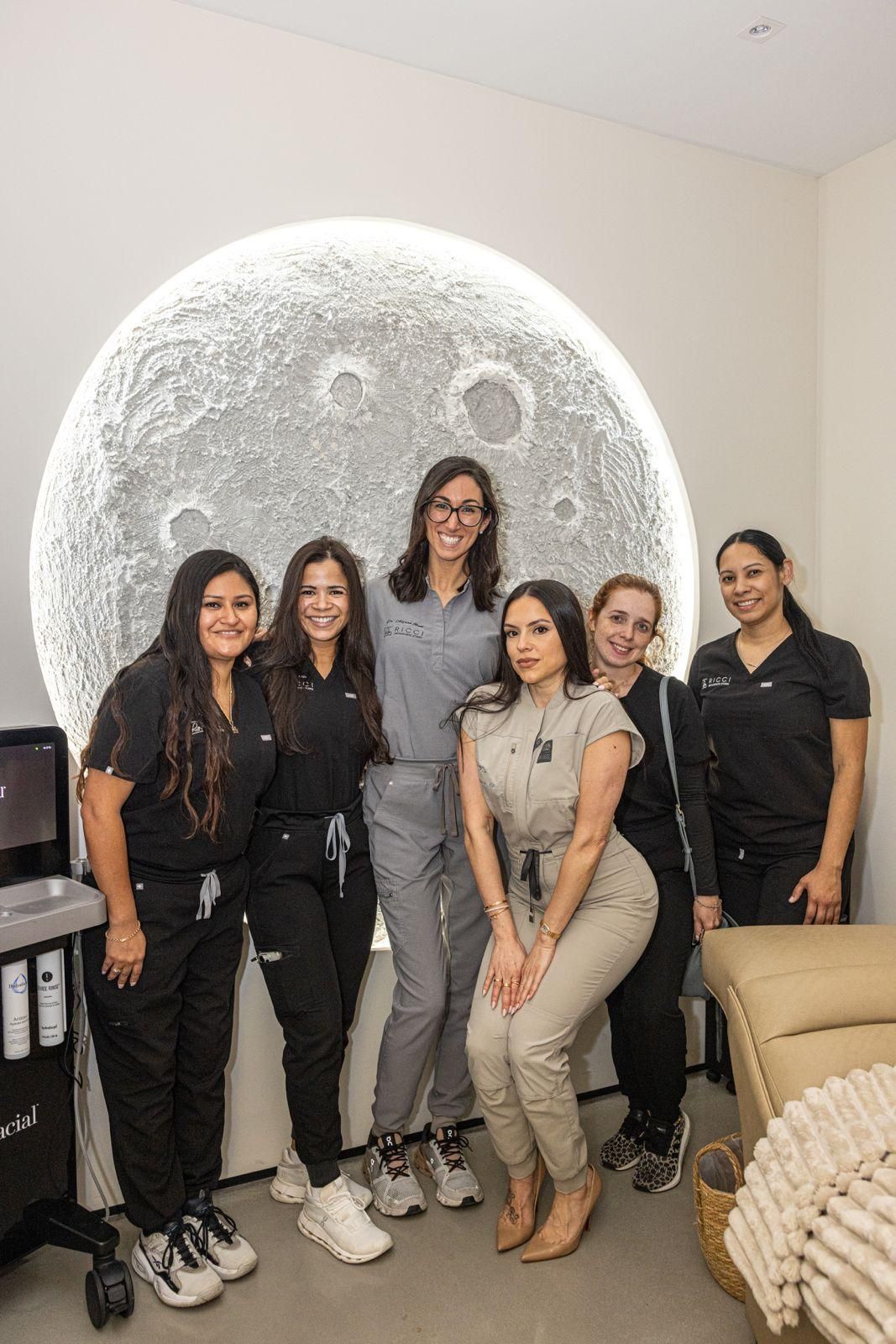 Six people pose in a row in a modern office with a large, illuminated circular moon decoration on the wall.