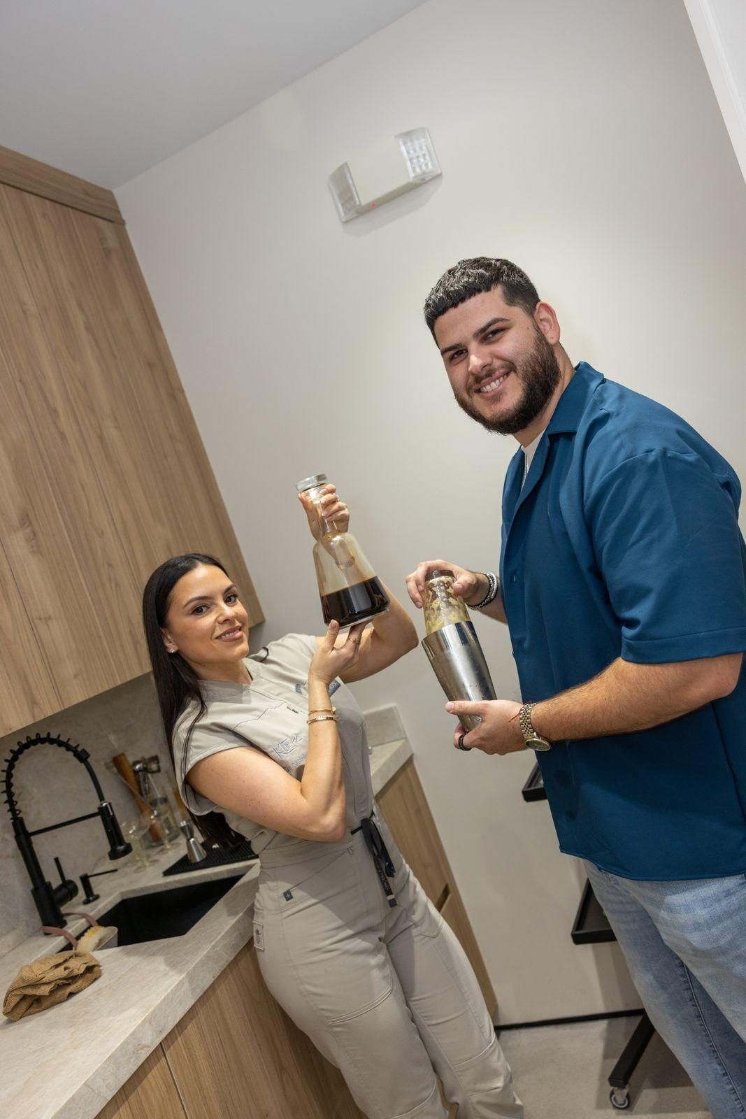 Two people smiling in a modern kitchen; one holds a glass carafe of coffee, the other holds a cocktail shaker.