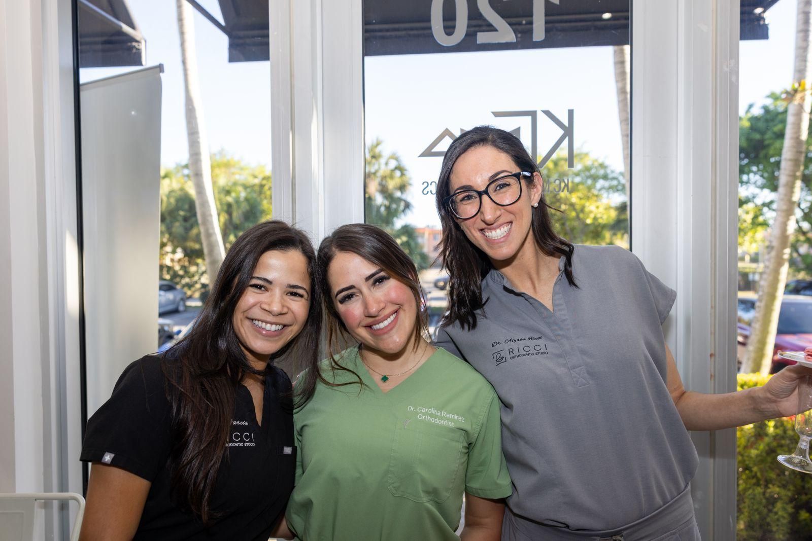 Three smiling colleagues in scrubs pose together indoors in front of a windowed storefront.
