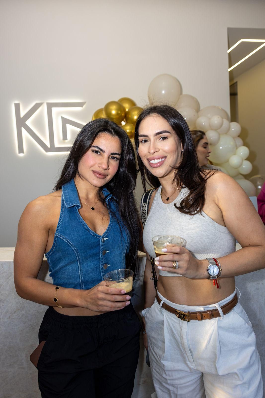 Two people posing and smiling indoors, holding drinks, in front of a white wall with gold balloons and a logo.
