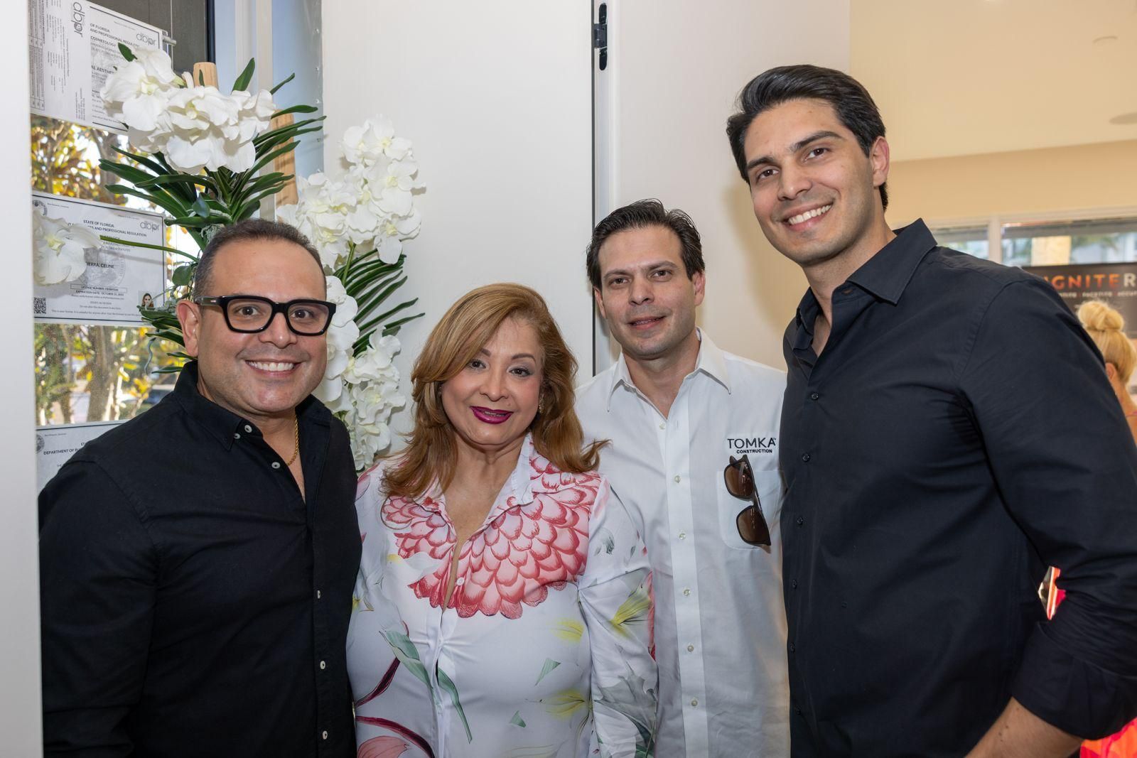 Four people pose for a photo at an indoor event, standing together and smiling in front of white floral arrangements.